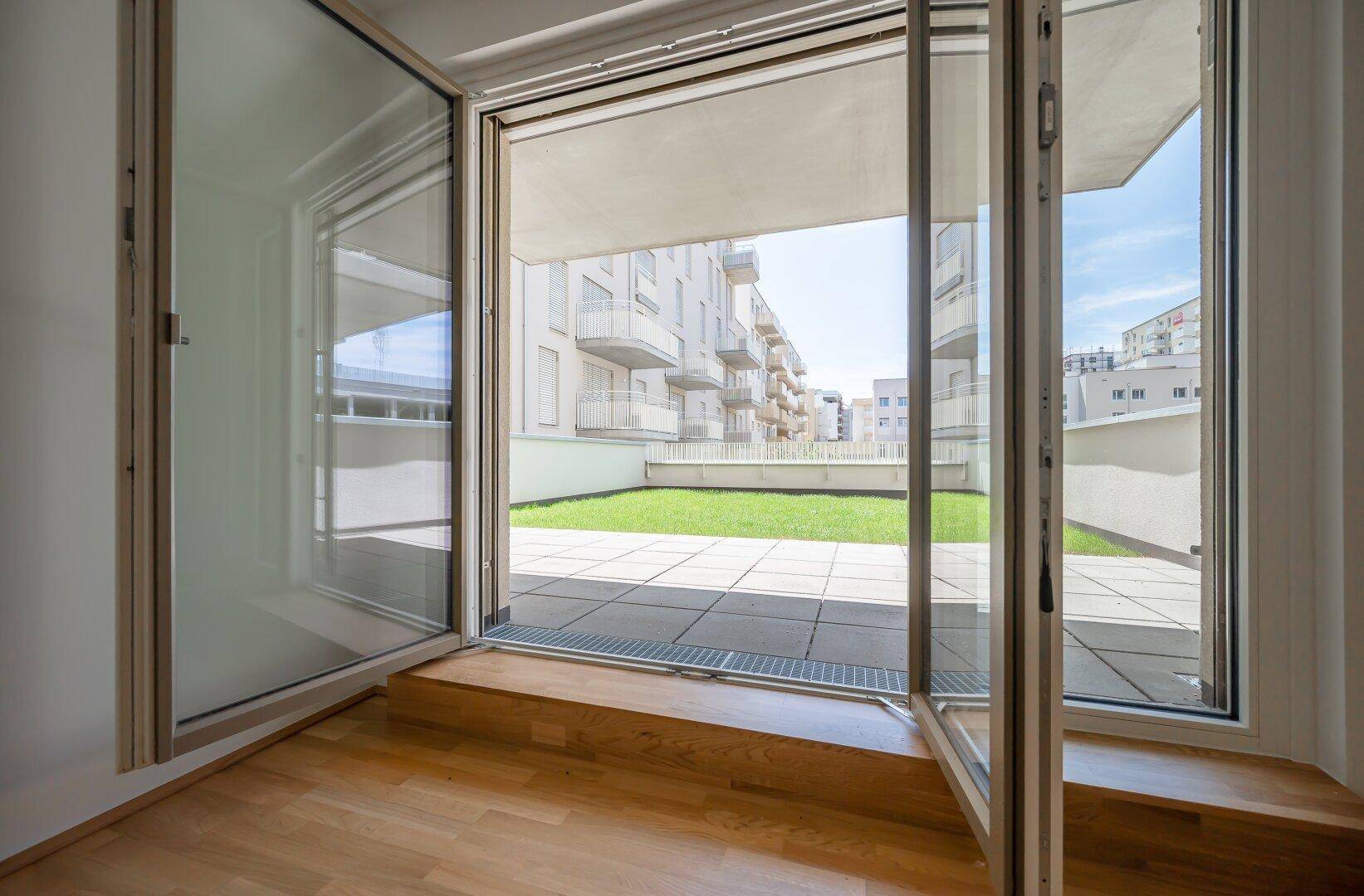 Blick aus dem Innenraum durch große Glastüren auf die grüne Terrasse.