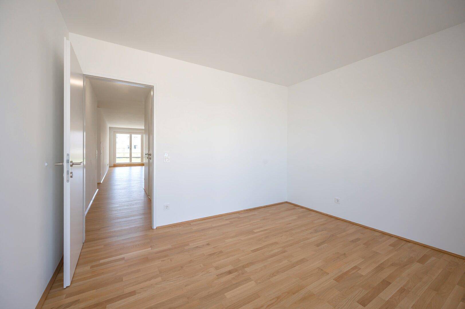Leeres Zimmer mit Parkettboden und offener Tür zum hellen Flur.
