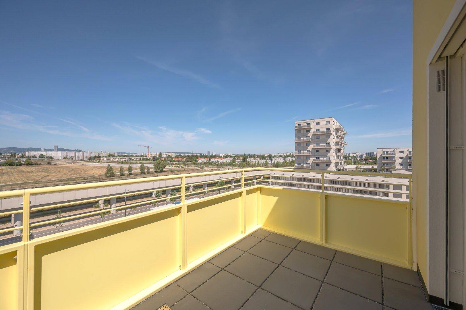 Sonniger Balkon mit gelbem Geländer und weitem Blick auf die Stadt und umliegende Gebäude.