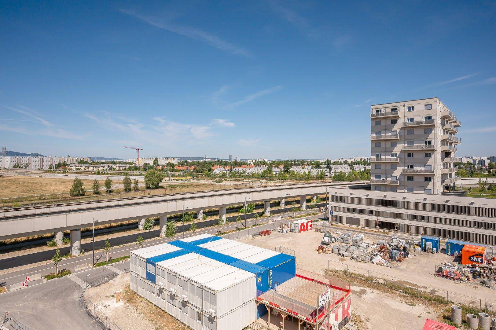 Weitläufige Ansicht einer Baustelle mit umliegenden Gebäuden und einer Bahntrasse im Hintergrund.