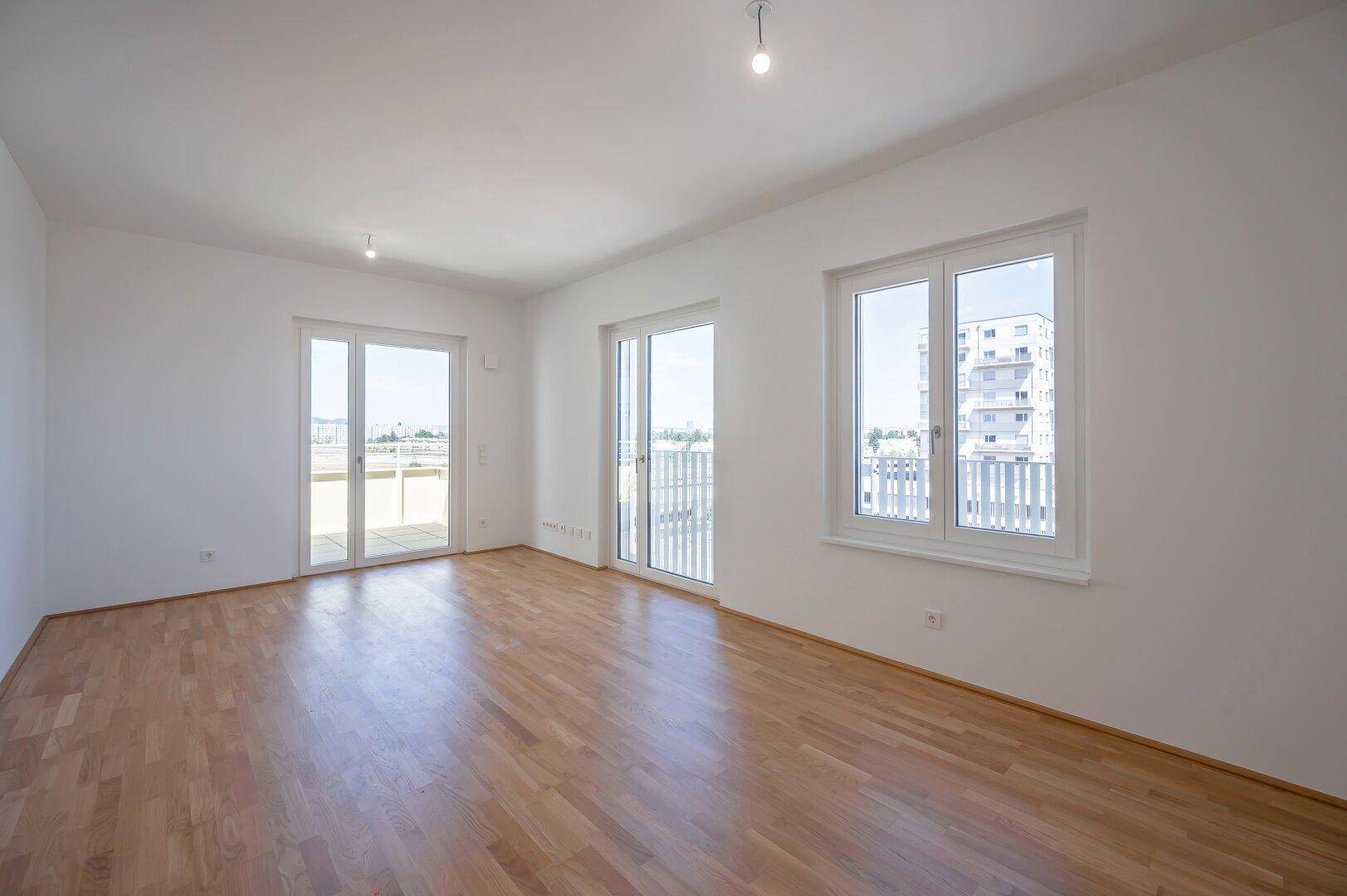 Helles, leeres Wohnzimmer mit hellem Parkettboden und Zugang zu einem Balkon.