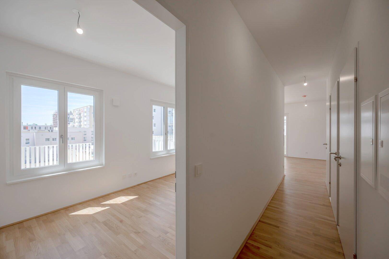 Heller Flur mit Holzfußboden, der in ein weiteres Zimmer mit großen Fenstern führt.