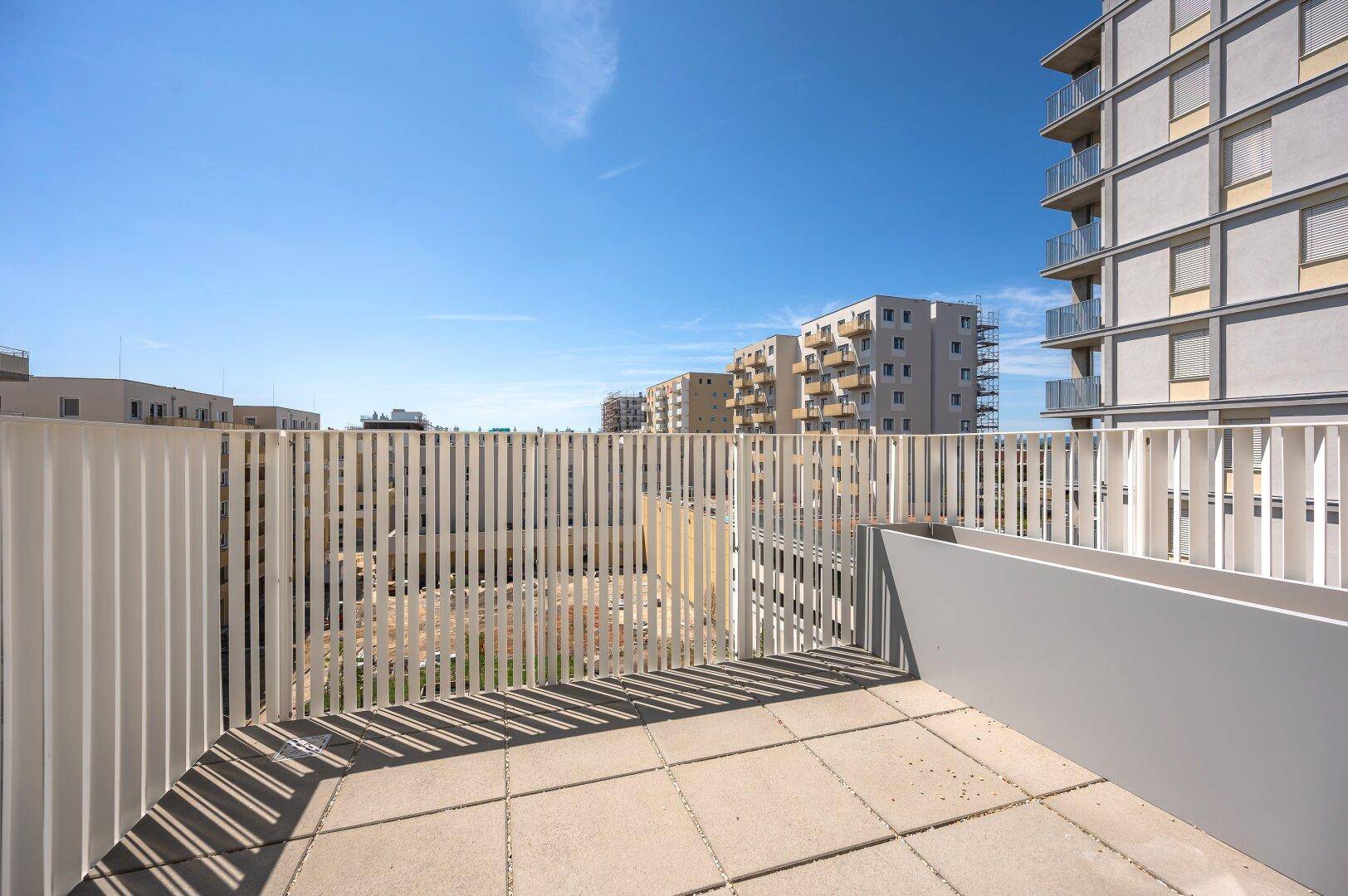 Sonnige Terrasse mit Fliesenboden und modernem Geländer, umgeben von Neubauten.