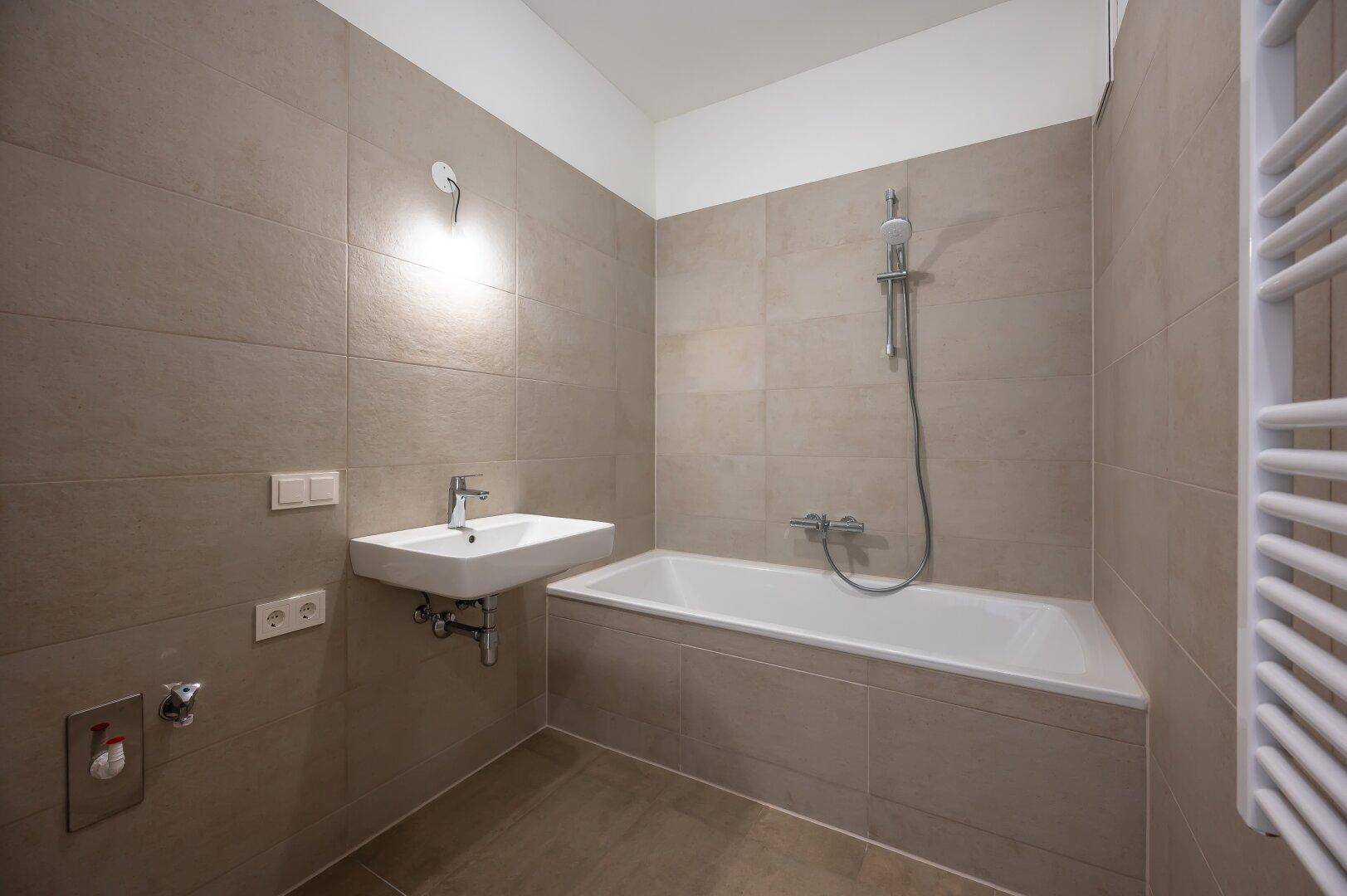 Modernes Badezimmer mit Badewanne, Waschbecken und beheiztem Handtuchhalter in hellen Fliesen.