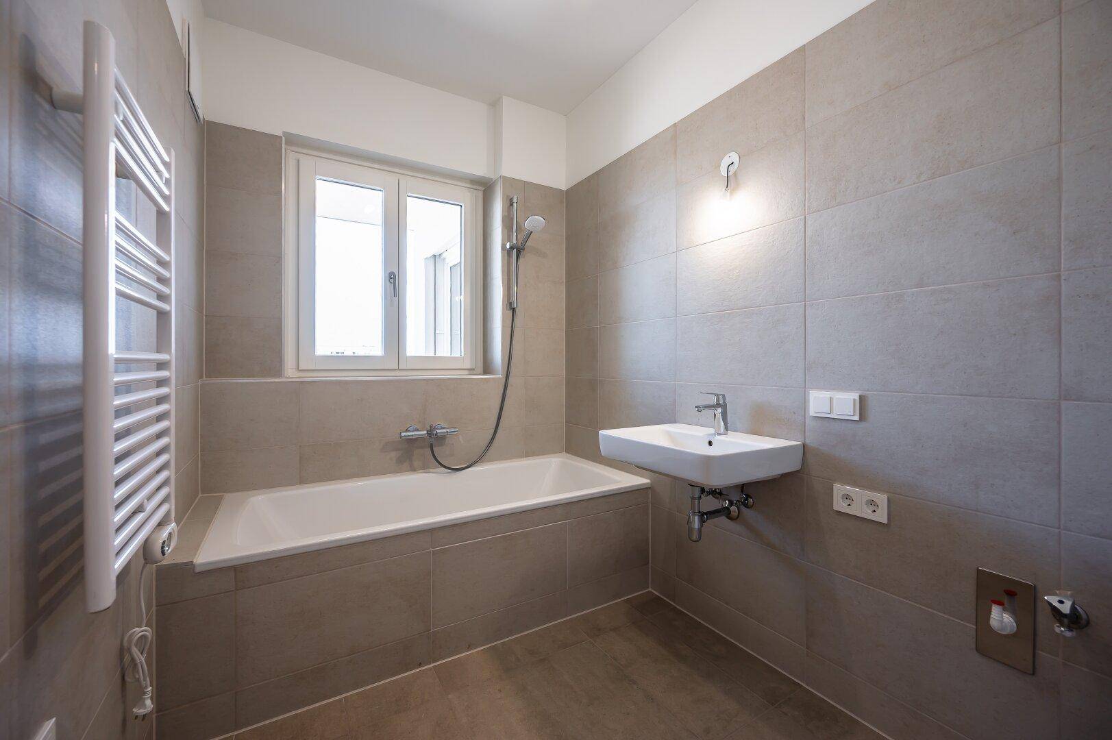 Helles Badezimmer mit Badewanne, Waschbecken, beheiztem Handtuchhalter und Fenster.