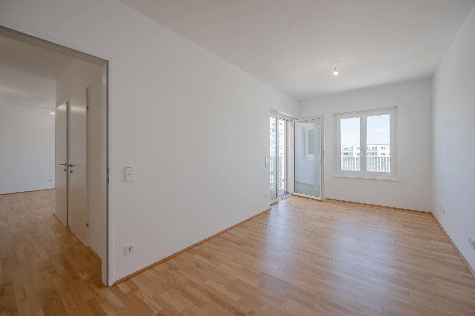 Geräumiges, unmöbliertes Wohnzimmer mit Parkettboden und Fensterfront zum Balkon.