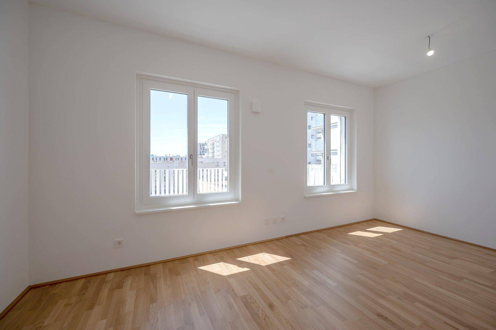 Helles Schlafzimmer mit Holzfußboden und zwei großen Fenstern, die viel Tageslicht hereinlassen.