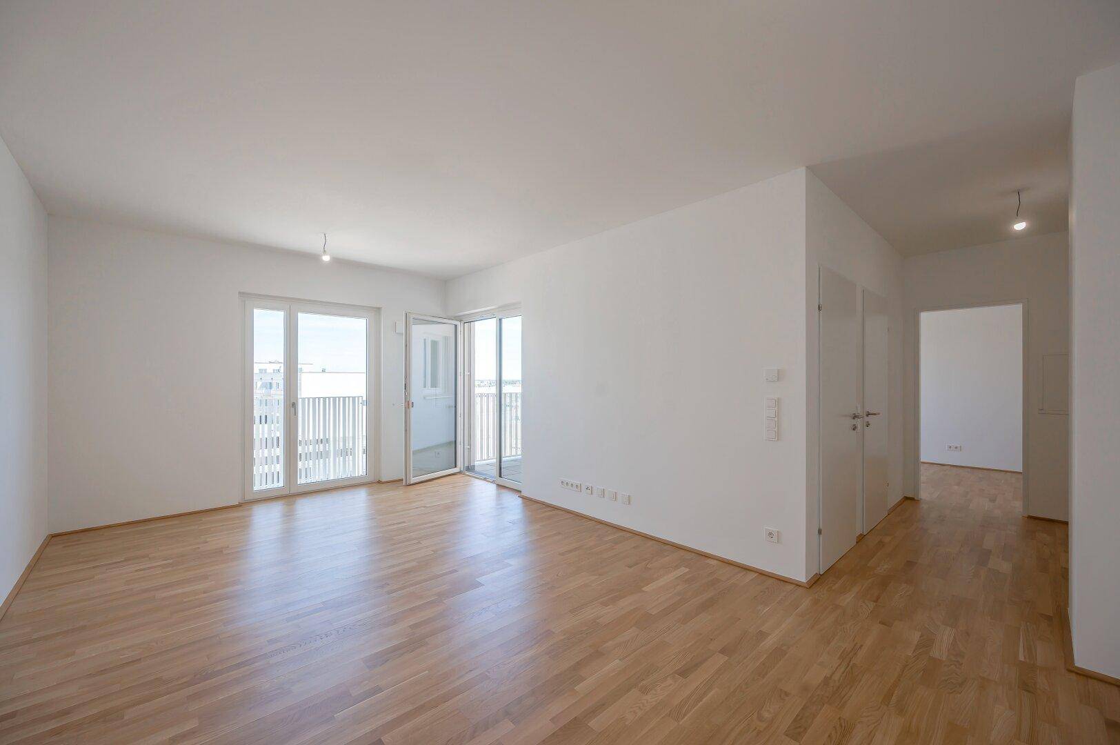 Geräumiges, leeres Wohnzimmer mit hellem Holzboden und direktem Zugang zu einem sonnigen Balkon.