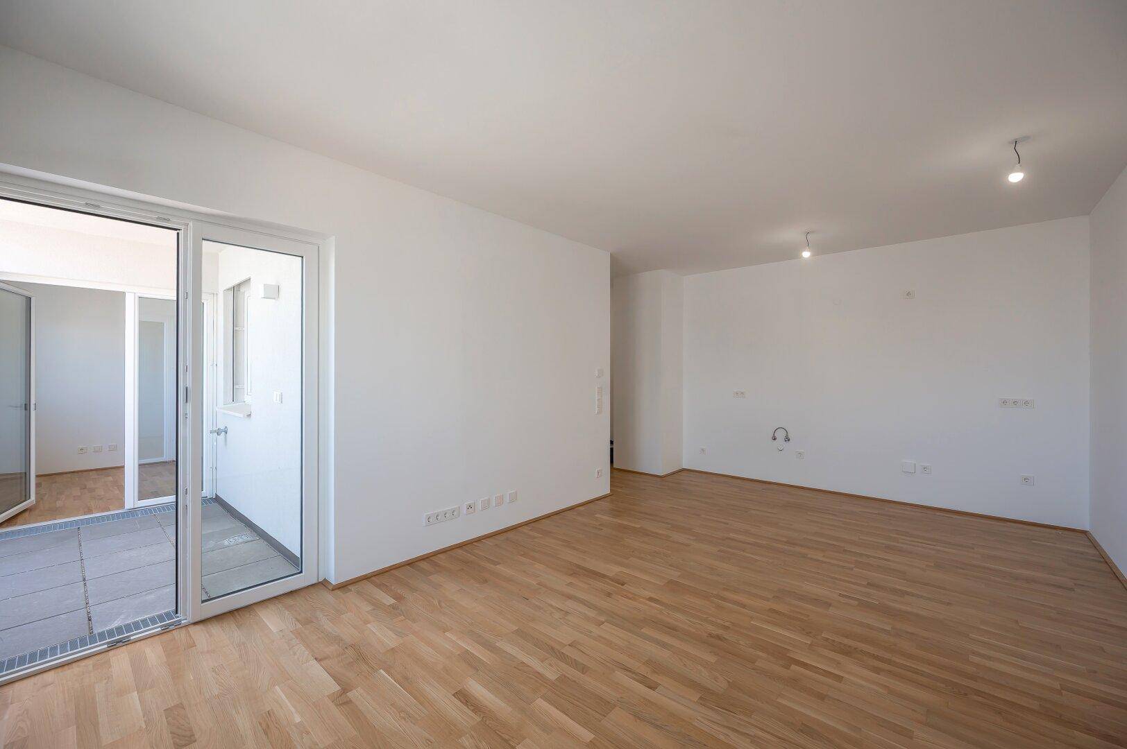 Geräumiger Wohnbereich mit Holzboden und breiter Schiebetür, die auf einen gefliesten Balkon führt.