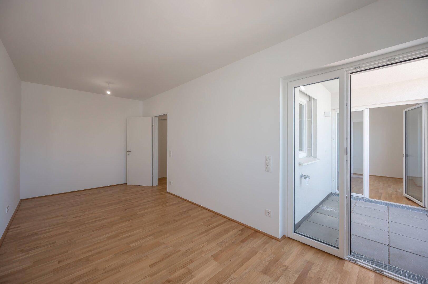 Geräumiger, leerer Raum mit Holzboden und direktem Zugang zu einem gefliesten Balkon.