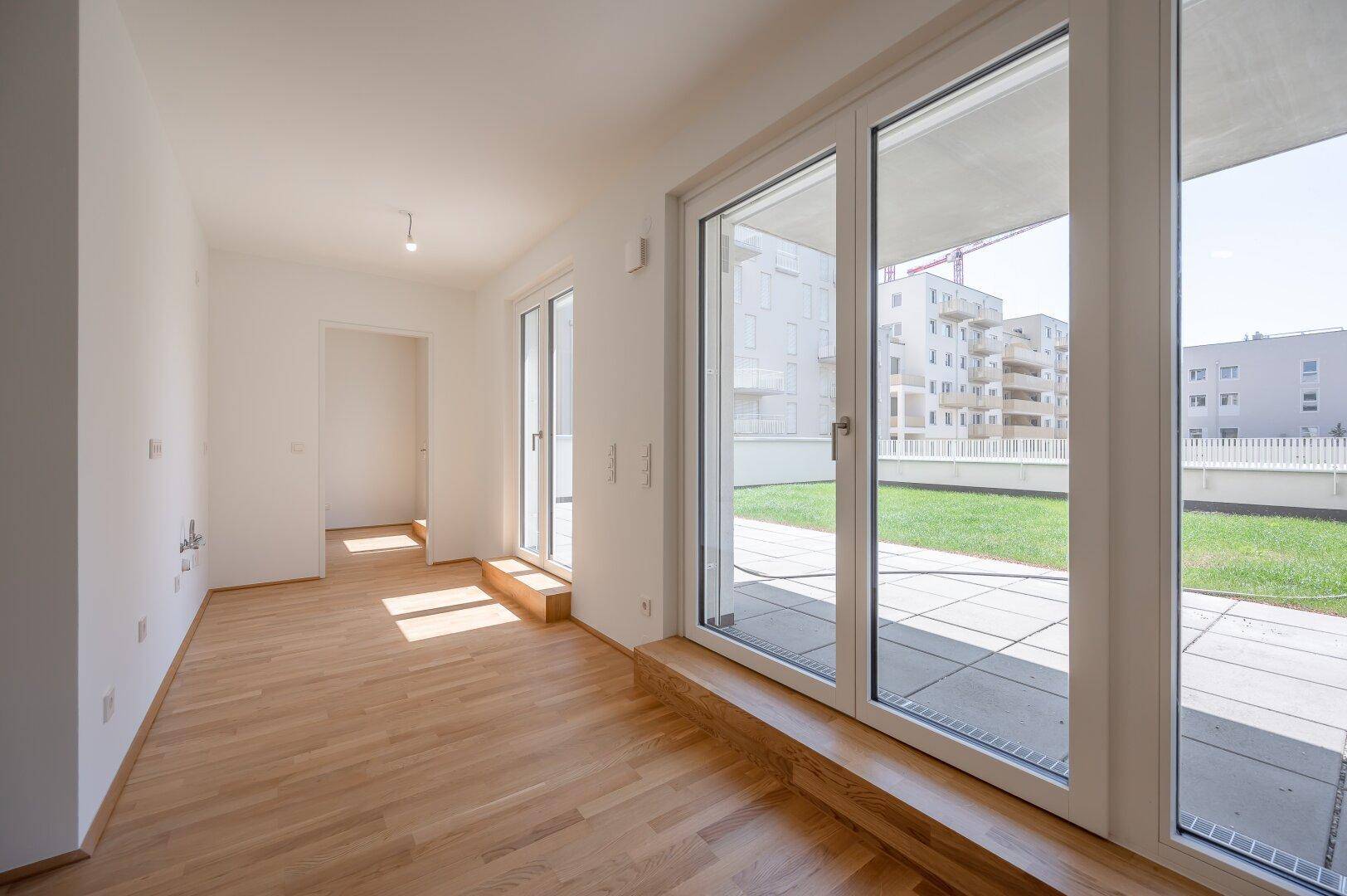 Heller, leerer Raum mit Holzboden und großen Fenstern zum Garten.