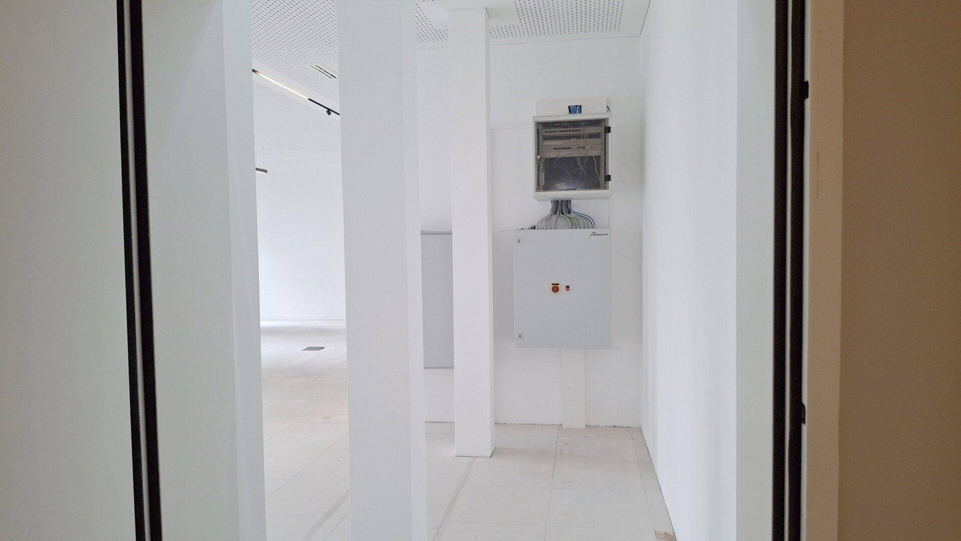 Leerer, heller Raum mit weißen Wänden, abgehängter Decke und sichtbaren Elektroinstallationen.