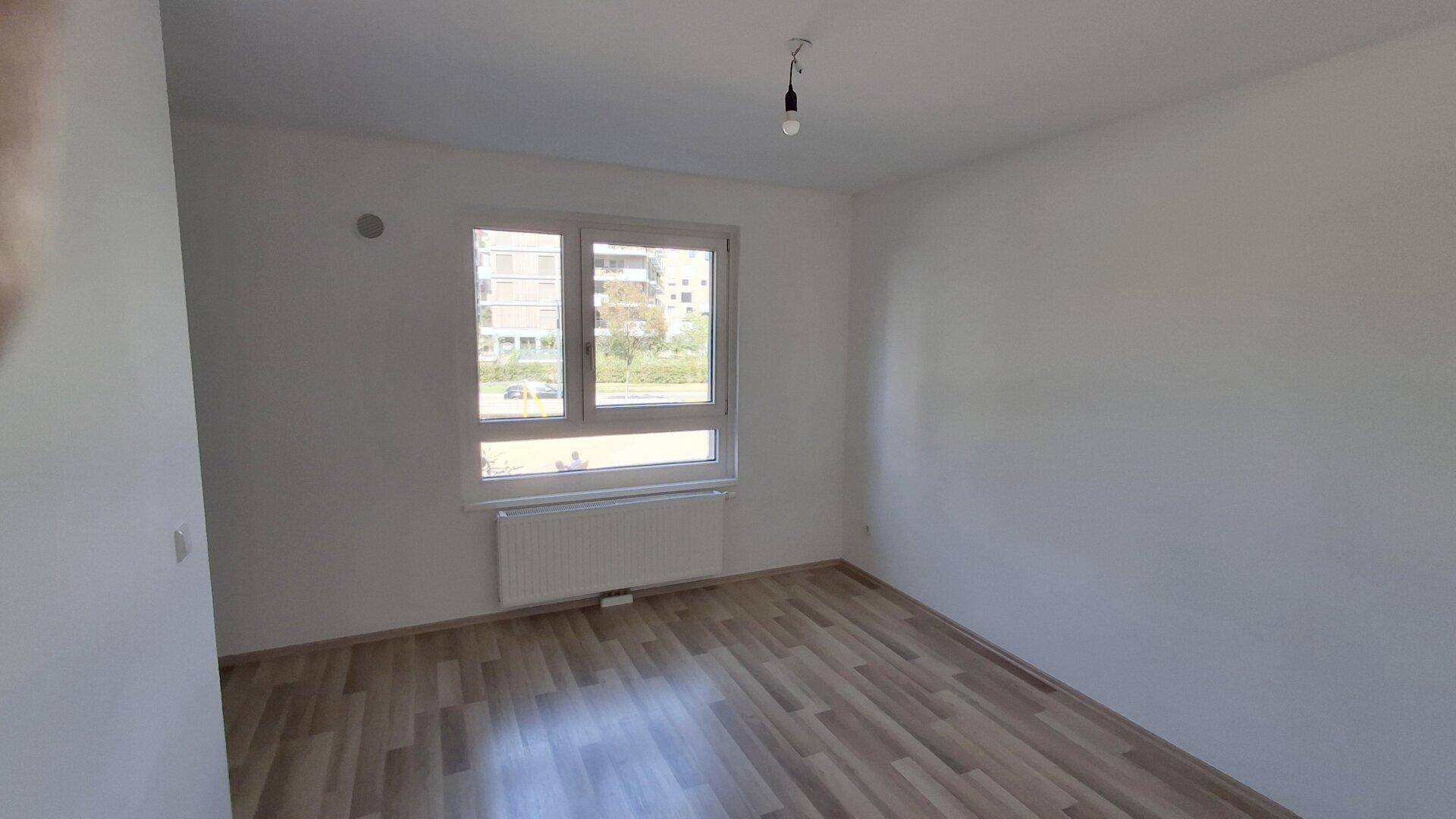 Leeres Schlafzimmer mit Laminatboden, großen Fenstern und Blick auf die umliegenden Gebäude.