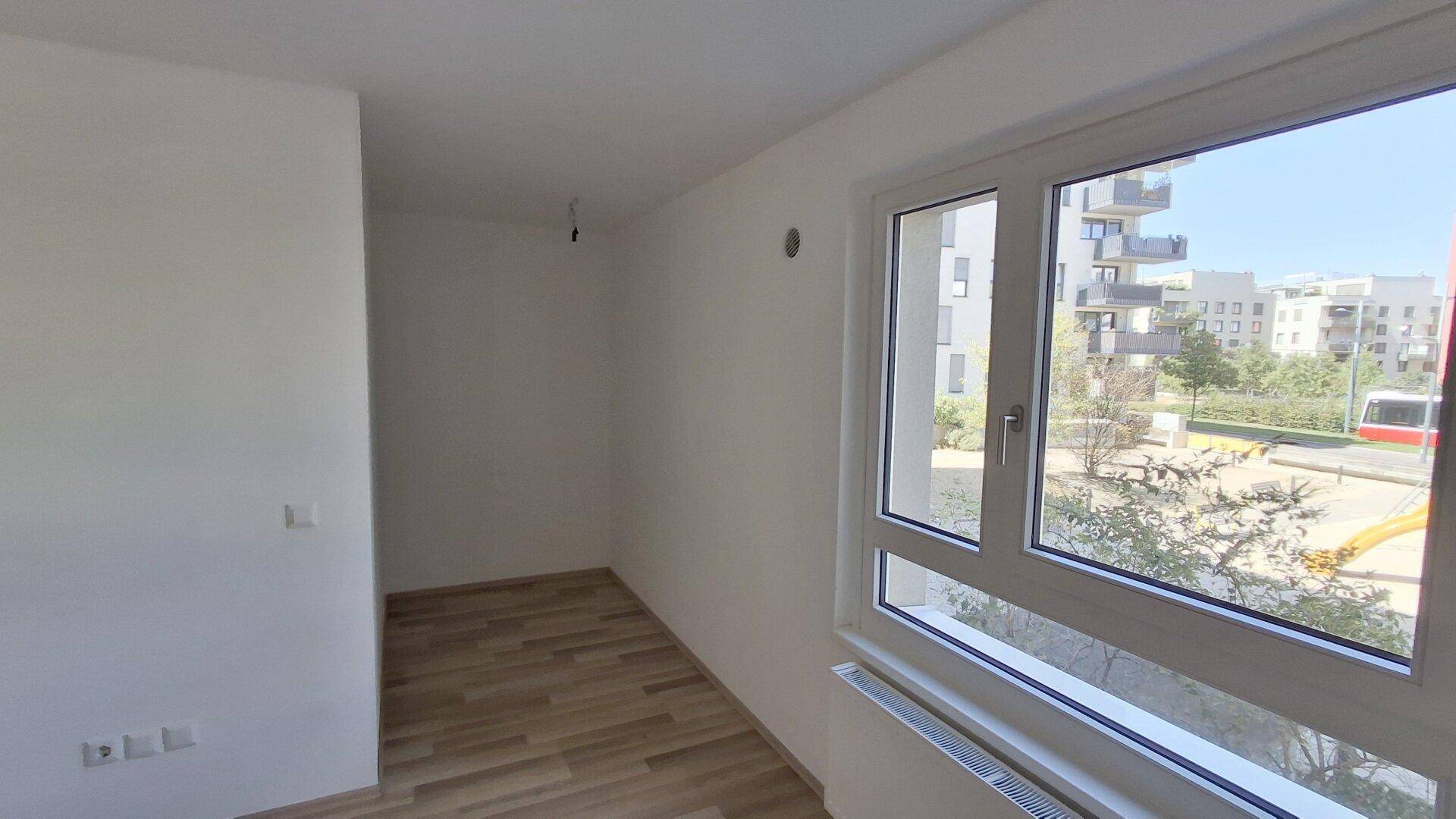 Helles Zimmer mit Laminatboden und Fenster, das einen Blick auf einen Spielplatz und Gebäude bietet.