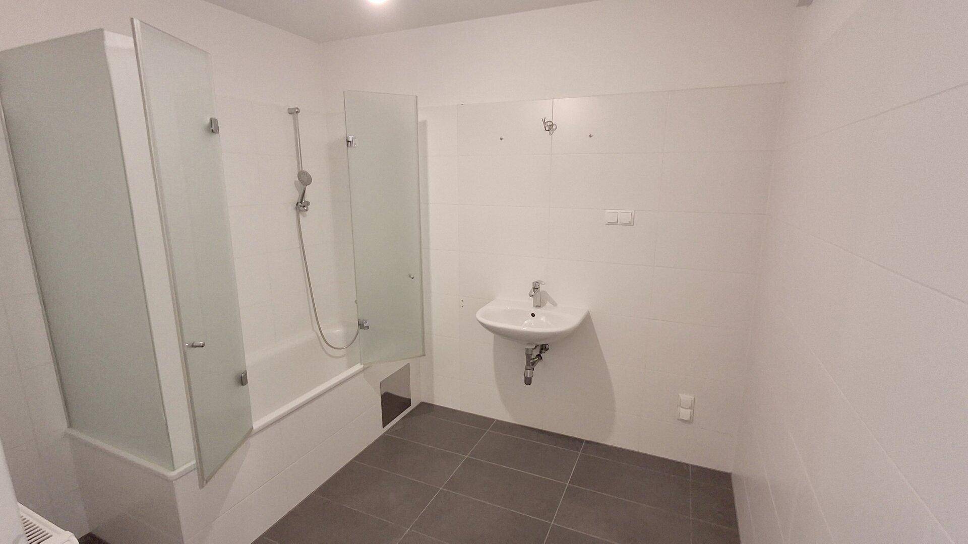 Modernes Badezimmer mit Badewanne, Duschabtrennung, Einzelwaschbecken und grauen Bodenfliesen.