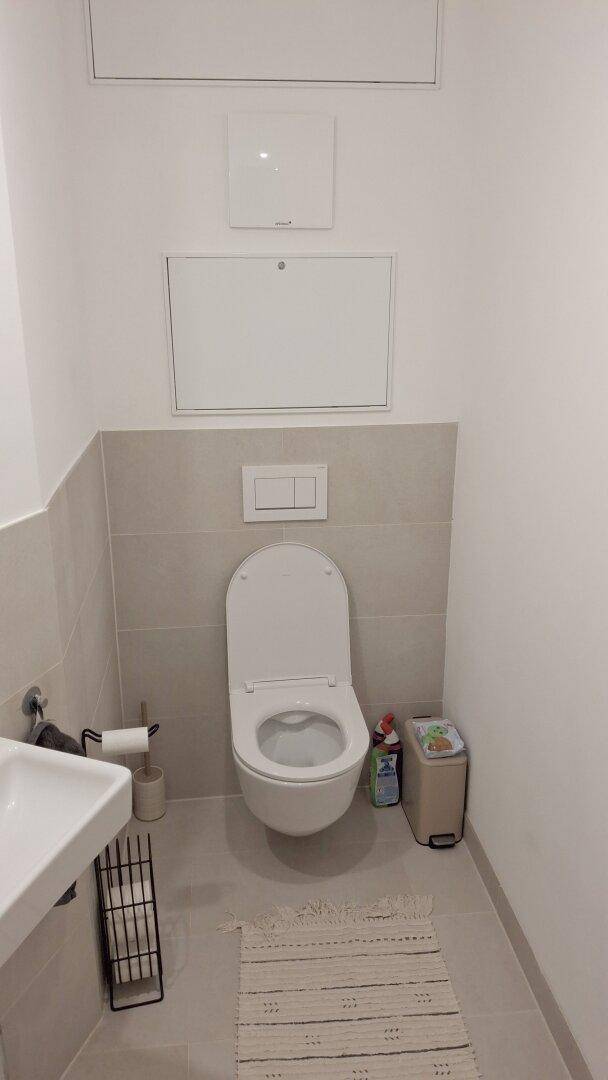 Separates WC mit wandhängender Toilette und hellgrauen Fliesen an der Wand.
