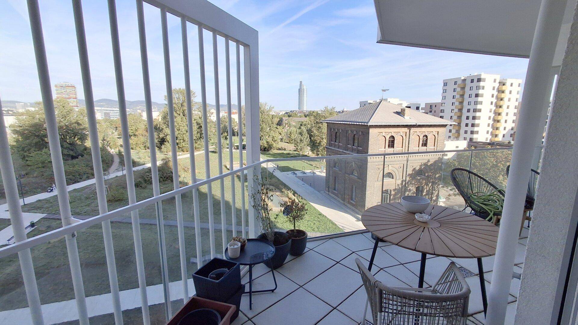 Möblierter Balkon mit Glasgeländer und weitem Blick über eine Parkanlage und die Stadt.