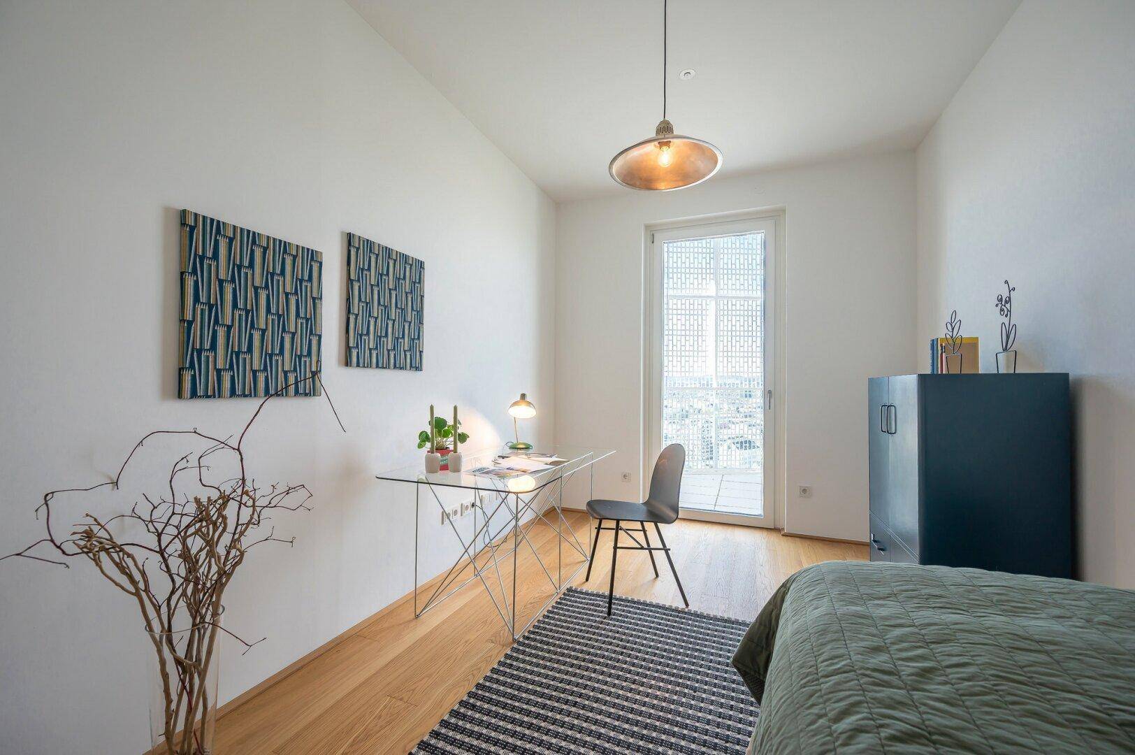 Helles Schlafzimmer mit Holzboden, Schreibtisch und Zugang zu einem kleinen Balkon.