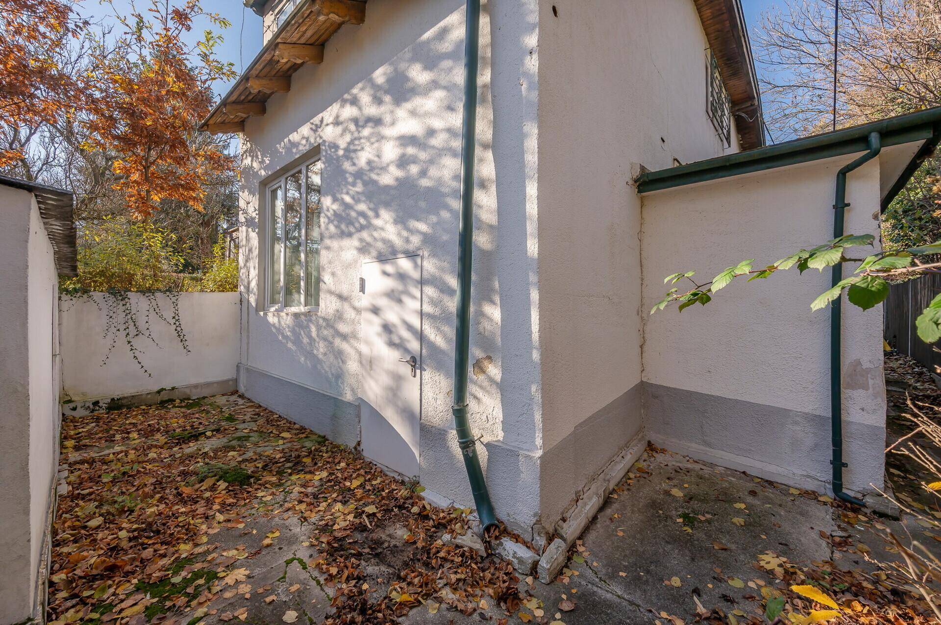 Seitenansicht des Hauses mit kleiner gepflasterter Fläche und herbstlichem Laub auf dem Boden.
