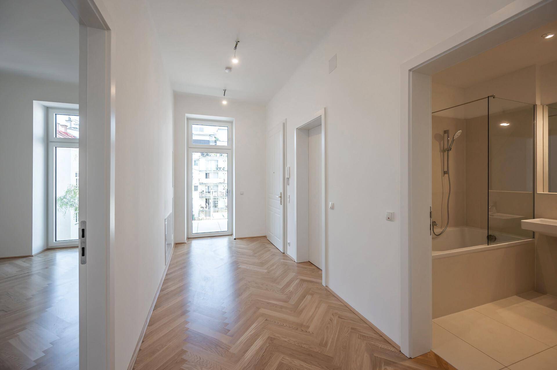 Heller Flur mit Fischgrätparkett, der zu einem Fenster führt, und Blick in ein modernes Badezimmer.