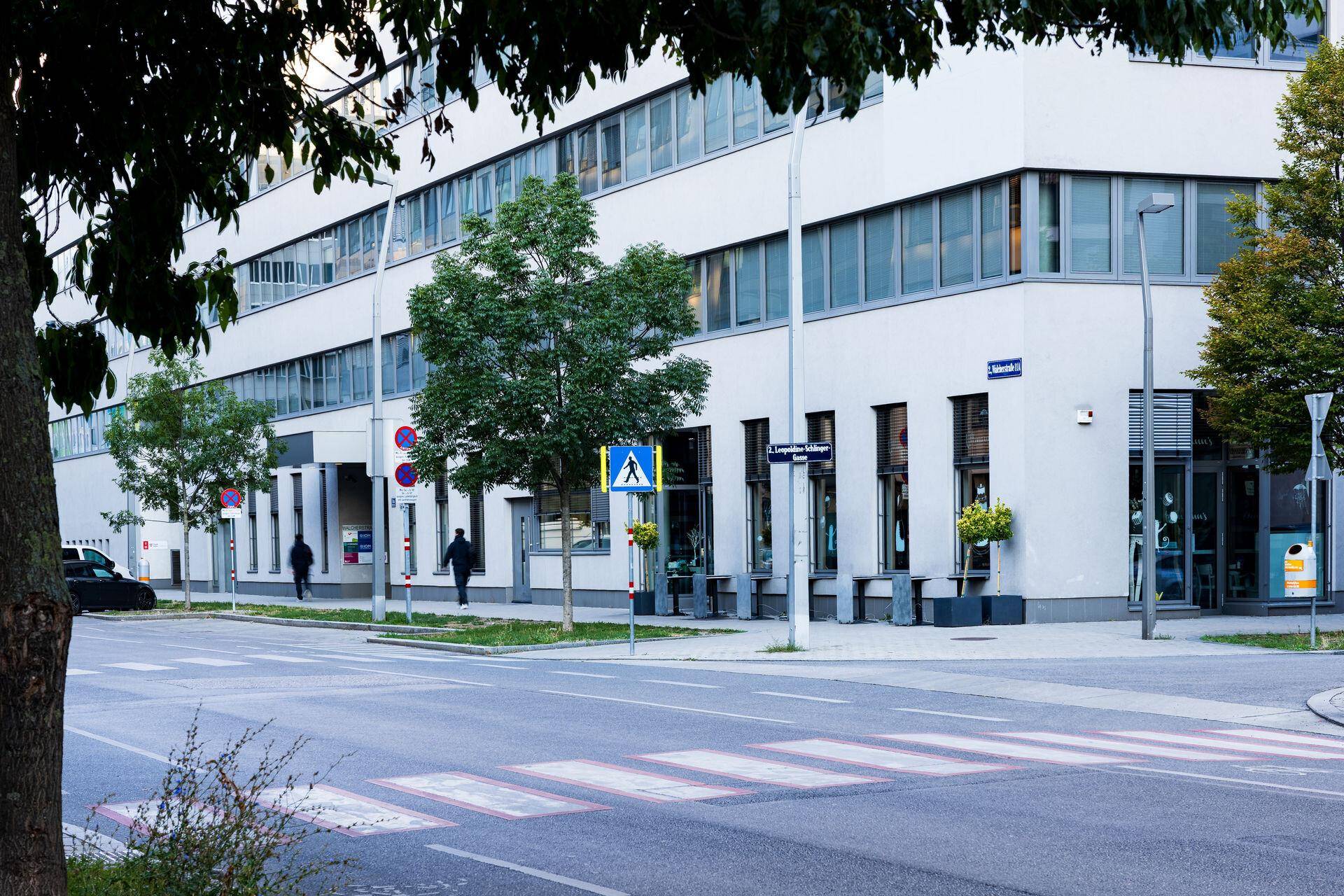 Straßenszene mit dem Erdgeschoss eines modernen Gebäudes, Geschäften und einem Zebrastreifen.