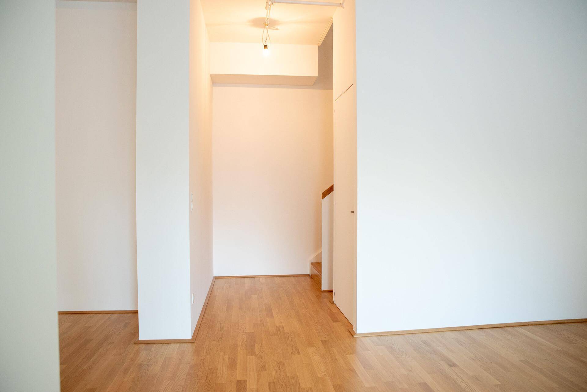 Heller Flur mit Holzboden und weiß gestrichenen Wänden, führt zu einem Treppenaufgang.