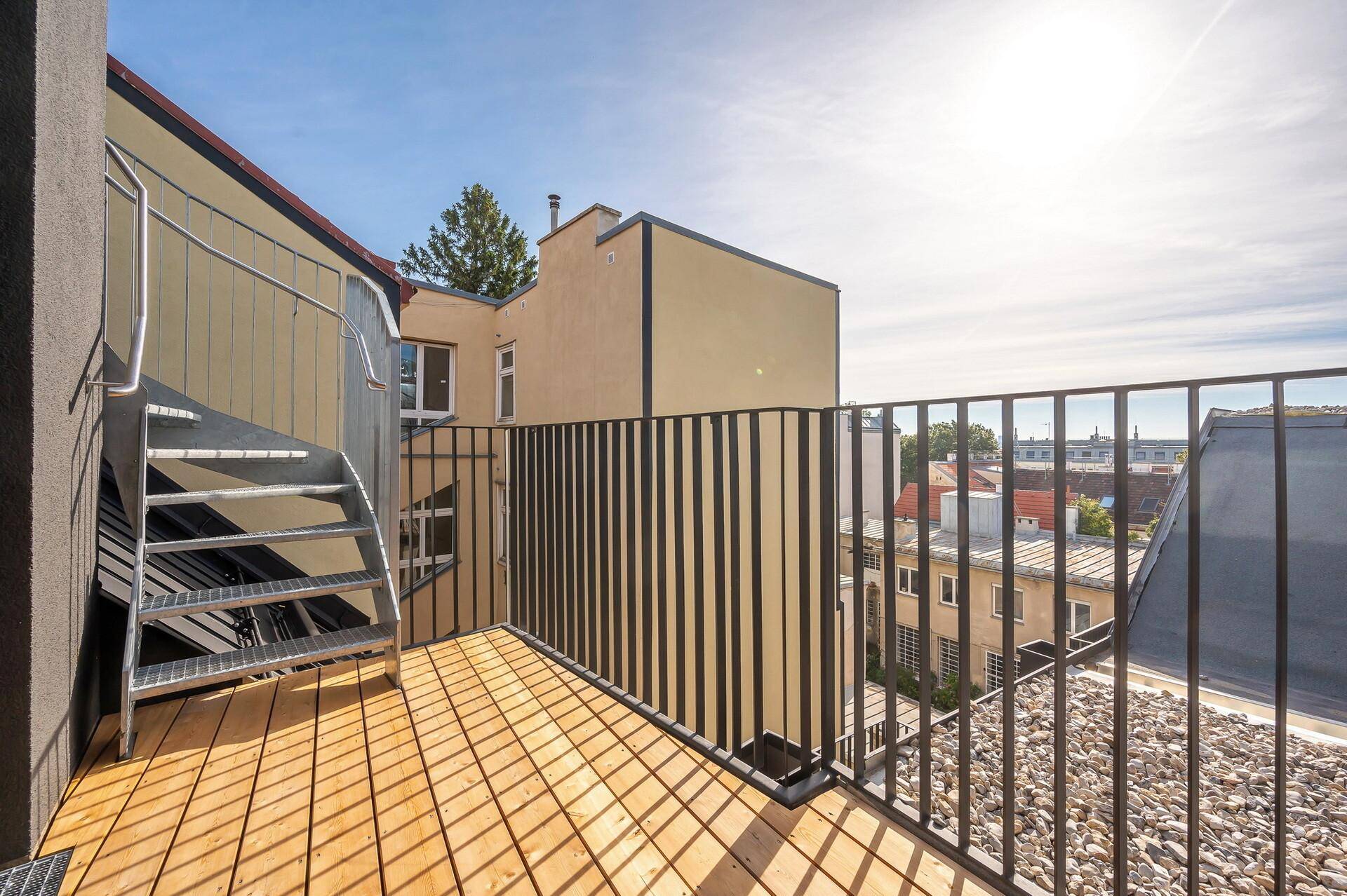 Balkon mit Holzbelag, Metallgeländer und einer Außentreppe, umgeben von Stadtgebäuden.