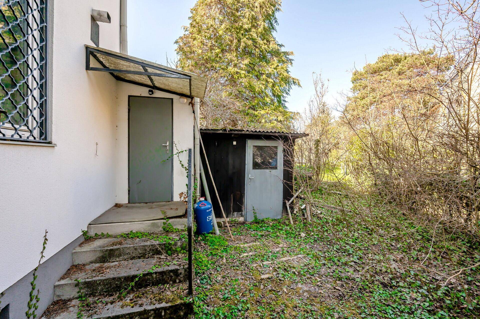 Hintereingang des Hauses mit Treppe und einem kleinen, rustikalen Gartenschuppen im Grünen.