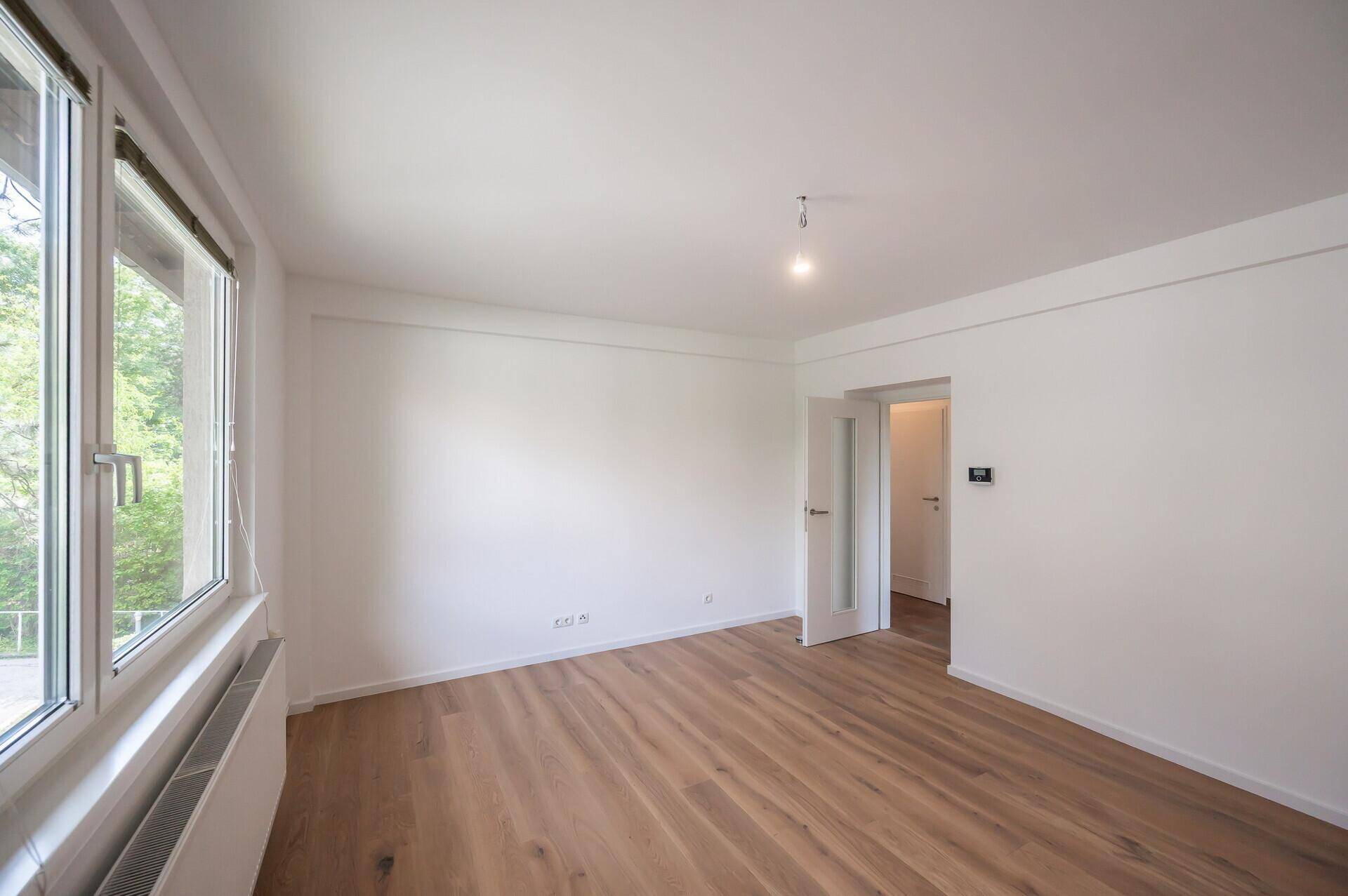 Helles Zimmer mit Holzboden und großen Fenstern, die viel Licht hereinlassen und eine Tür zeigen.