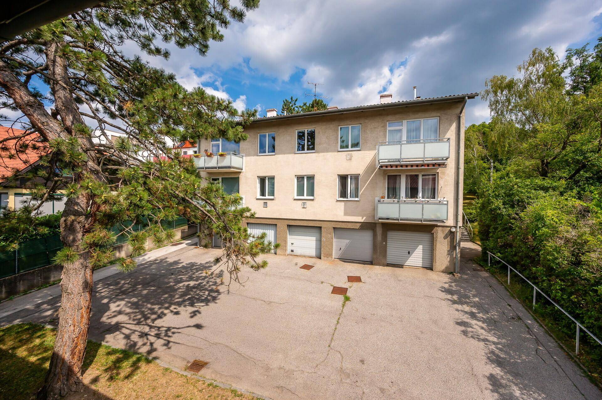 Rückansicht eines Mehrfamilienhauses mit Garagen und Balkonen, umgeben von viel Grün.