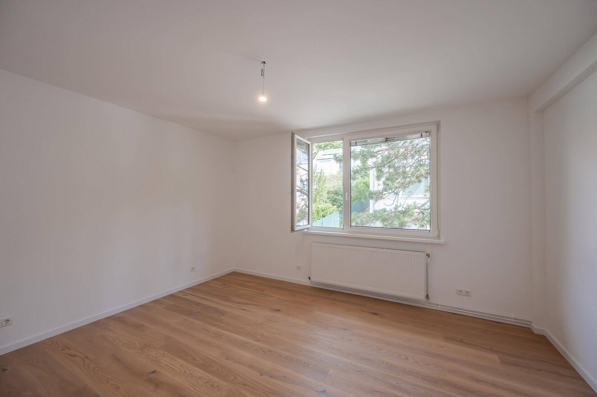 Geräumiges, unmöbliertes Zimmer mit Holzboden und einem Fenster, das den Blick ins Grüne freigibt.