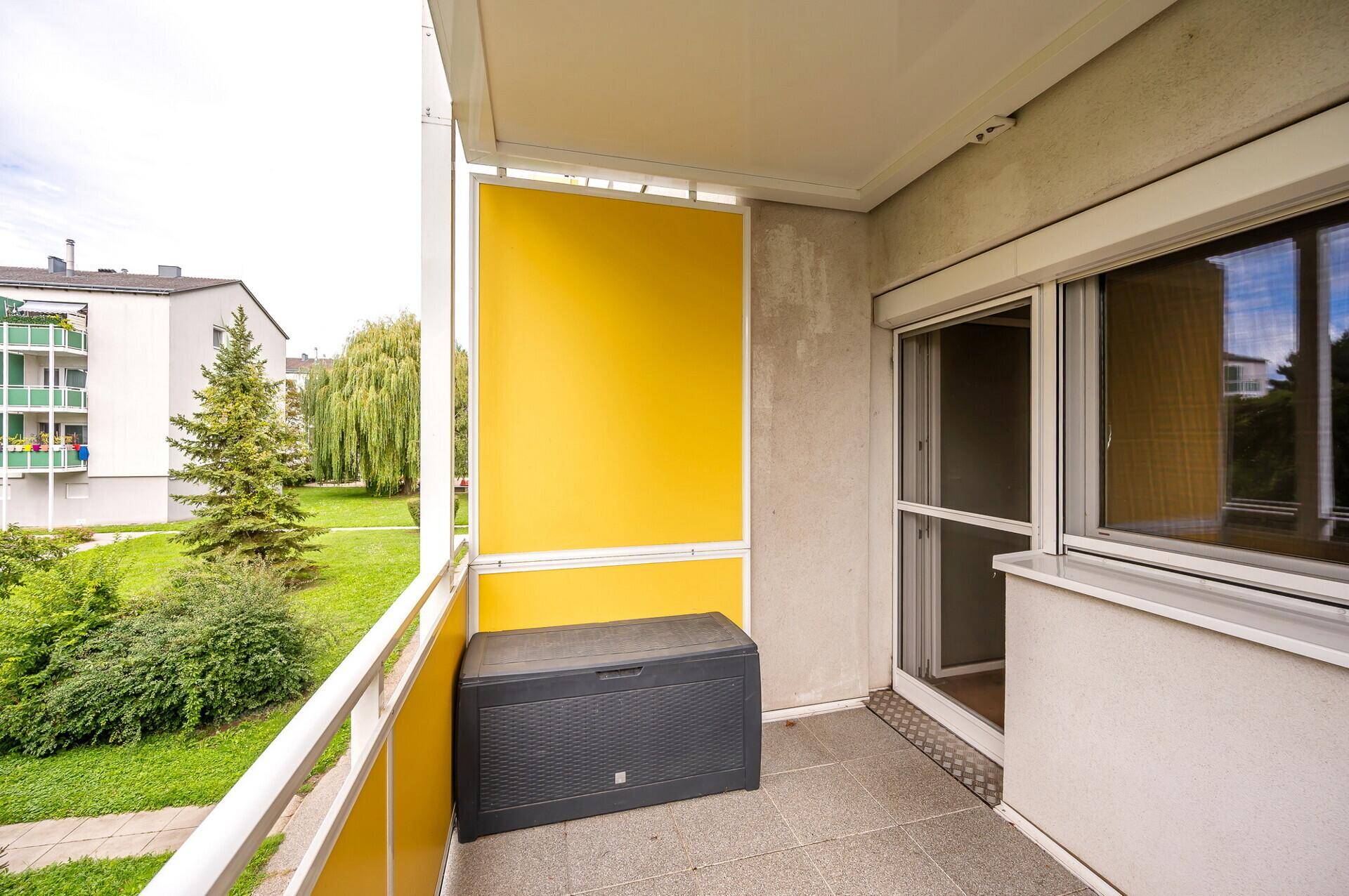 Heller Balkon mit gelbem Sichtschutz, Fliesenboden und Blick ins Grüne.