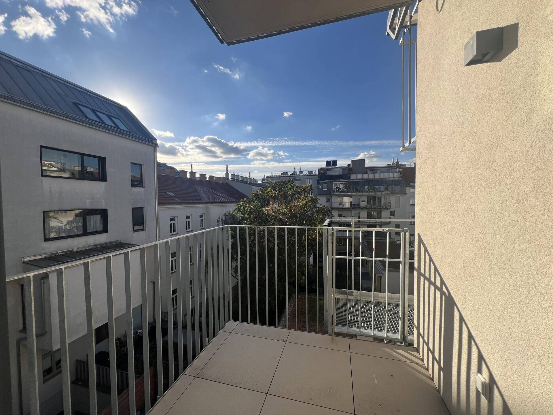 Heller Balkon mit Fliesenboden und Blick auf die umliegenden Gebäude und den blauen Himmel.