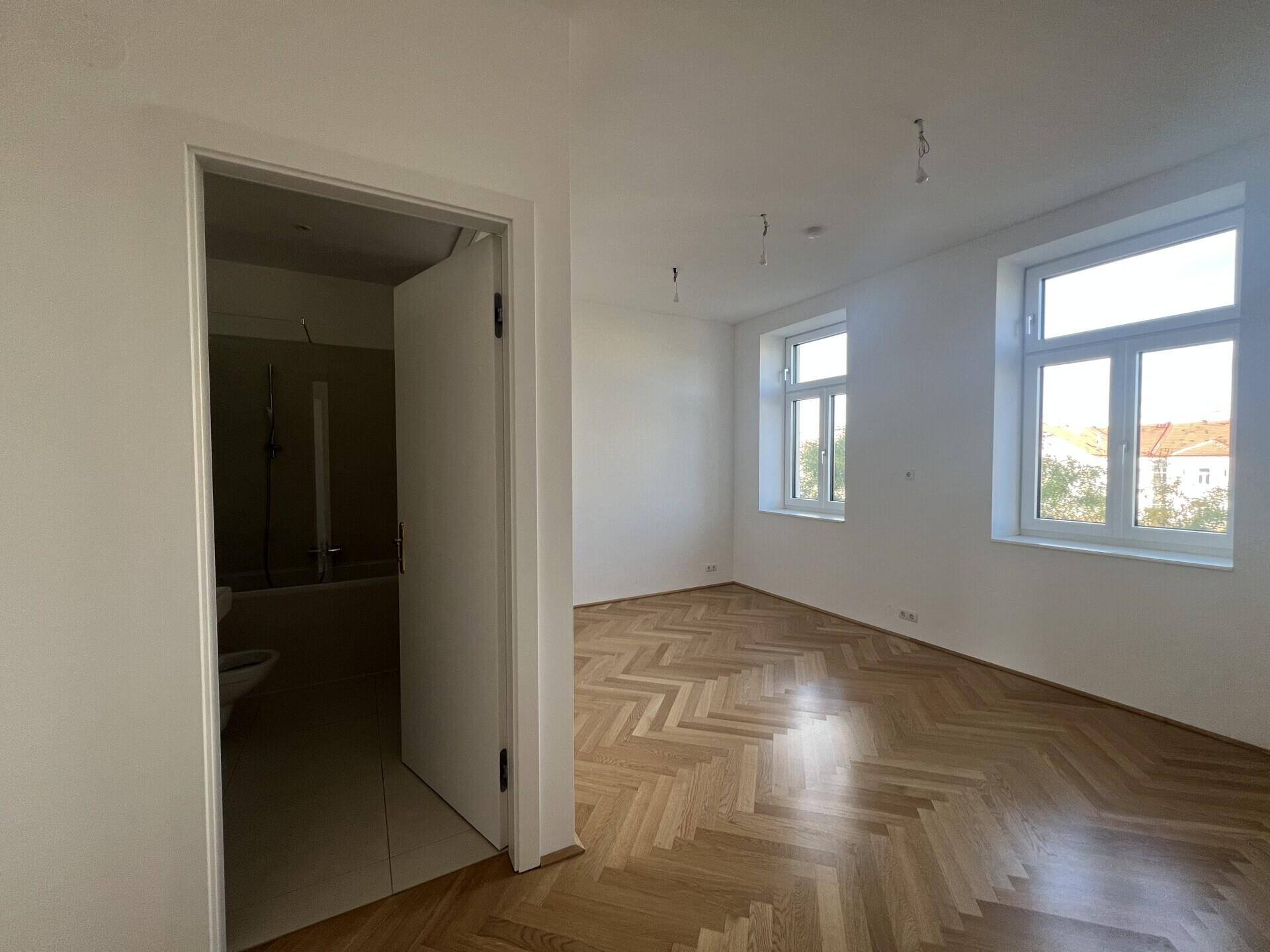 Blick aus einem leeren Zimmer mit Fischgrätparkett zum Eingang eines modernen Badezimmers.