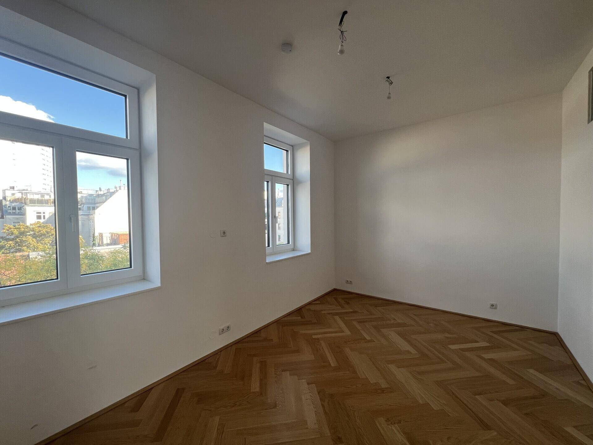 Leerer Raum mit Fischgrätparkett und zwei Fenstern, die viel Tageslicht hereinlassen.