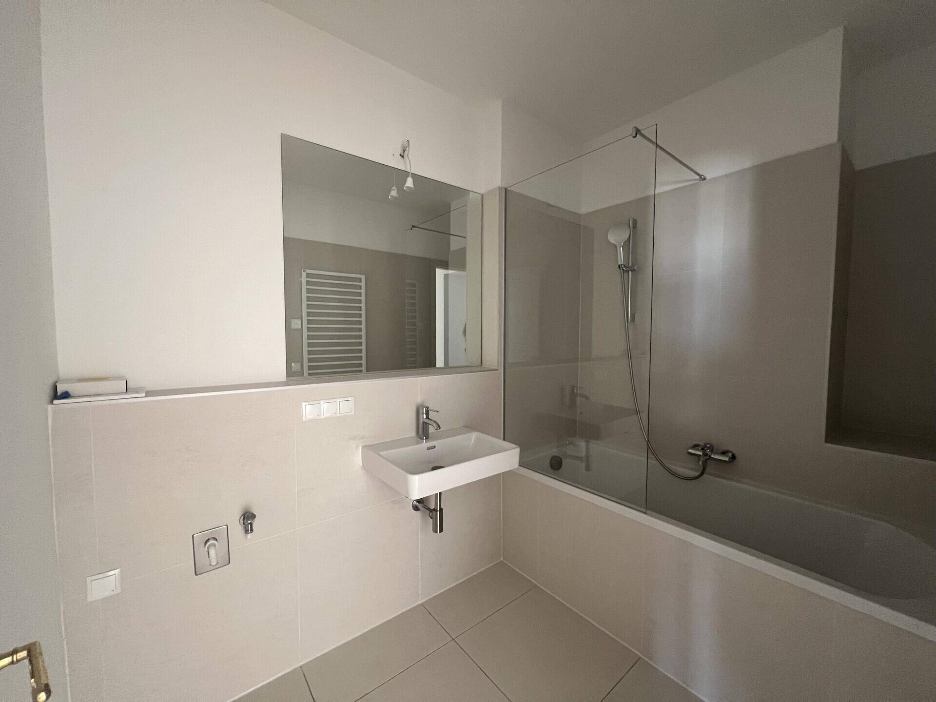 Modernes Badezimmer mit hellgrauen Fliesen, Badewanne mit Glasabtrennung und wandhängendem Waschbecken.
