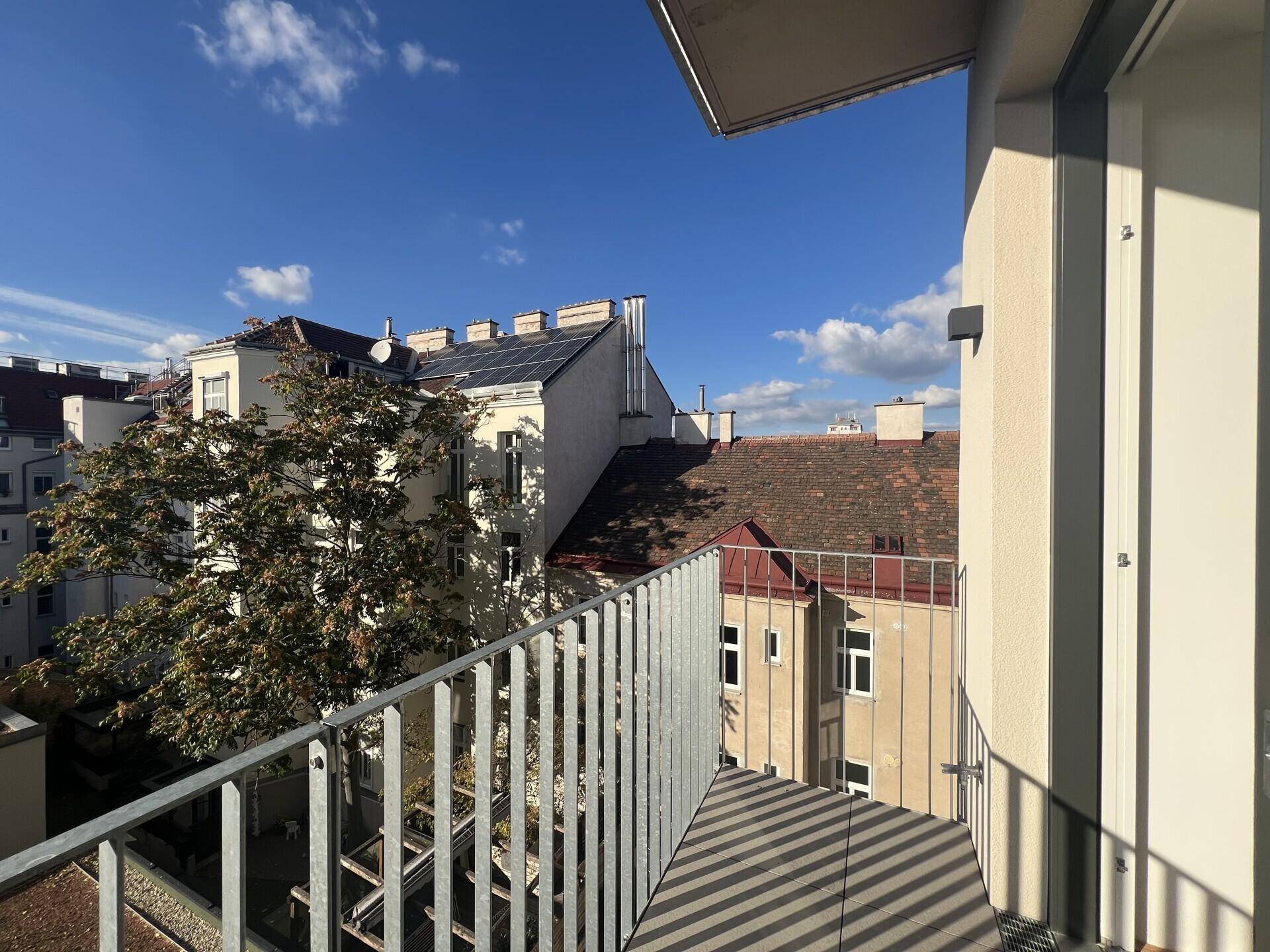 Blick vom Balkon auf die umliegenden Gebäude und die Straße, mit Bäumen und blauem Himmel.