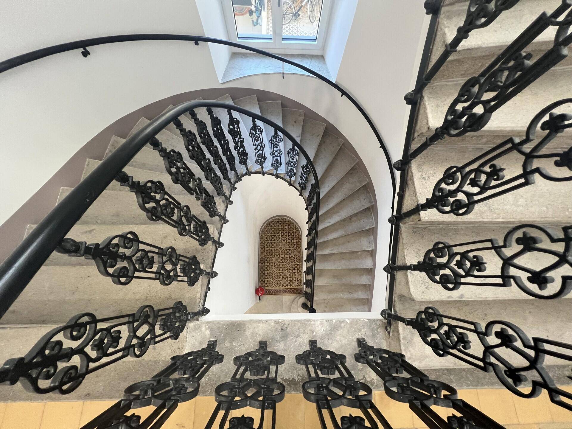 Blick von oben auf das spiralförmige Treppenhaus mit kunstvollen schmiedeeisernen Geländern und Steinstufen.