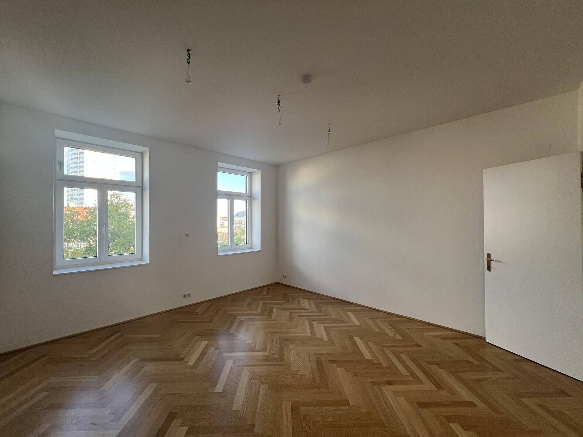 Geräumiges Zimmer mit Fischgrätparkett und zwei großen Fenstern, die viel Tageslicht hereinlassen.