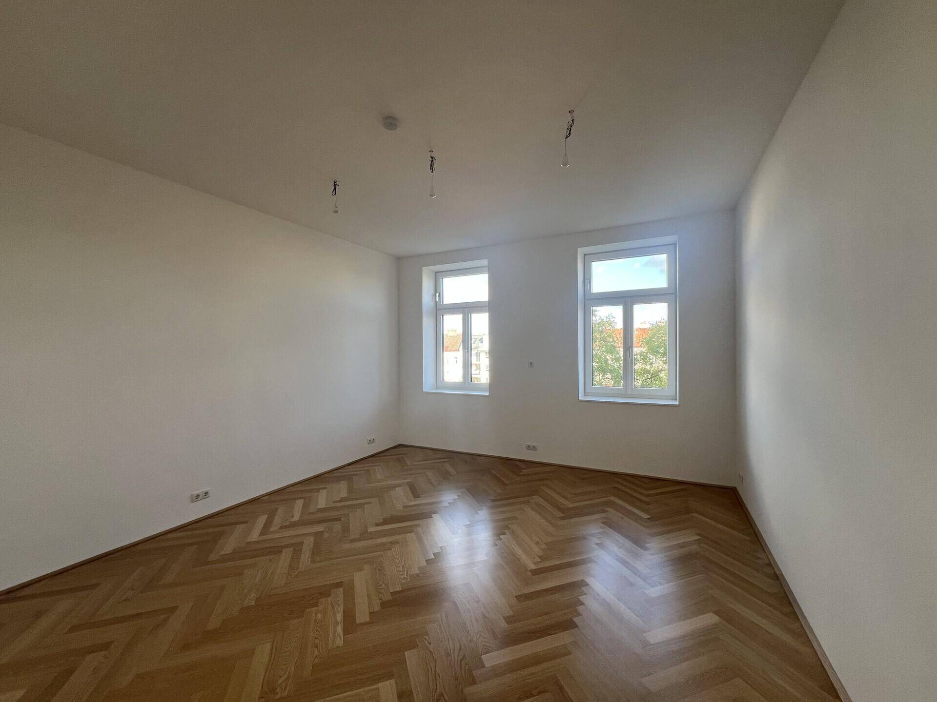 Helles Schlafzimmer mit zwei Fenstern und elegantem Fischgrätparkettboden, bereit zur Einrichtung.