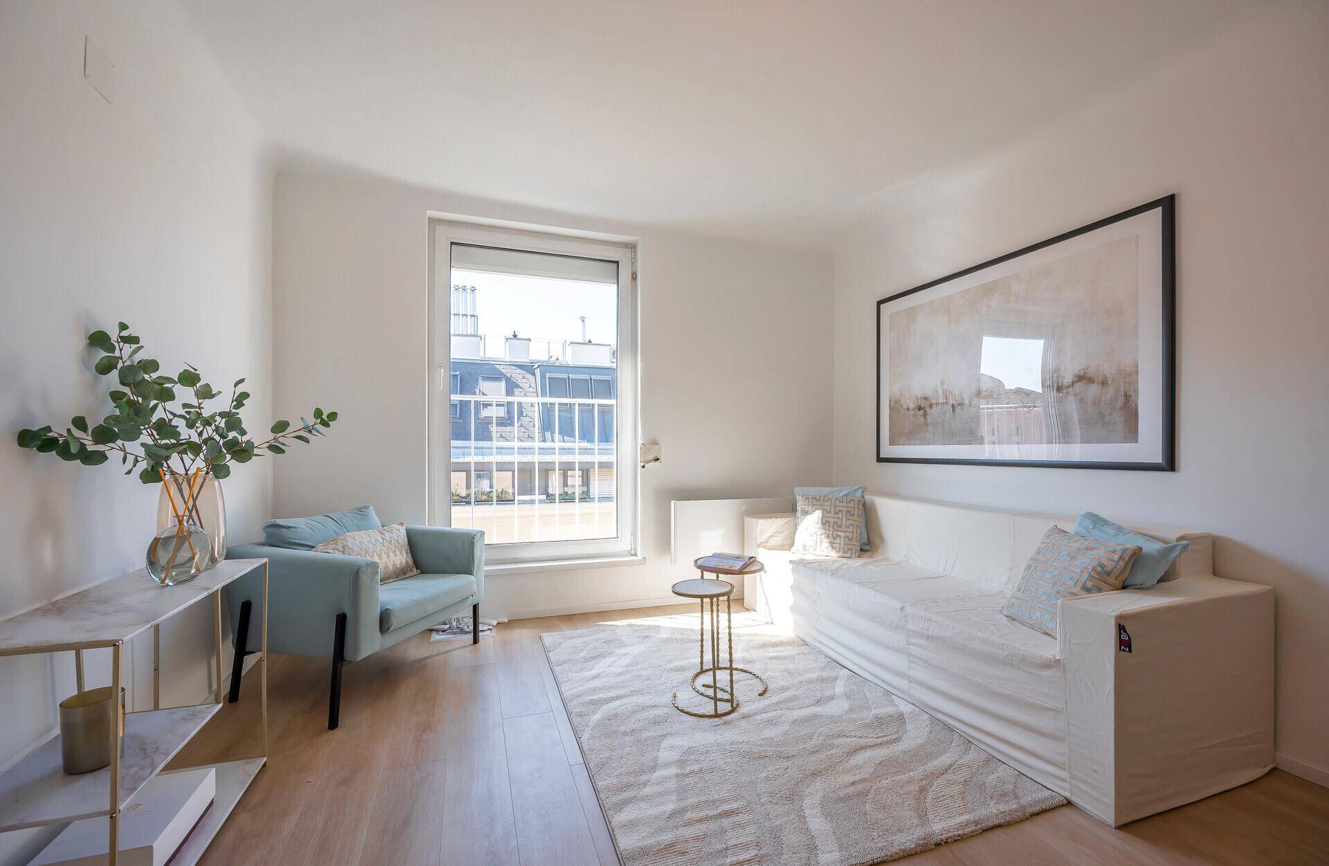 Gemütliches Wohnzimmer mit hellem Sofa, Sessel und Fenster mit Blick auf die umliegenden Gebäude.