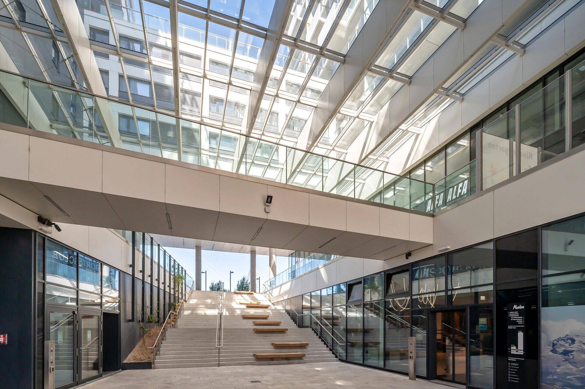 Heller, überdachter Bereich mit Glasdach, Treppen und Geschäften in einem modernen Gebäudekomplex.