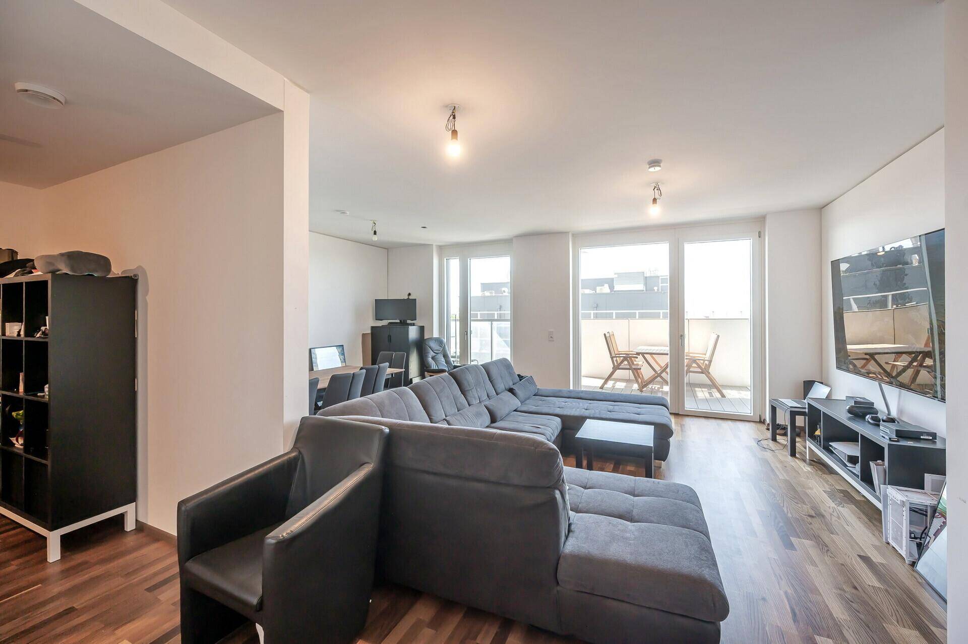 Geräumiges Wohnzimmer mit großem Ecksofa, Holzboden und Zugang zu einem sonnigen Balkon.