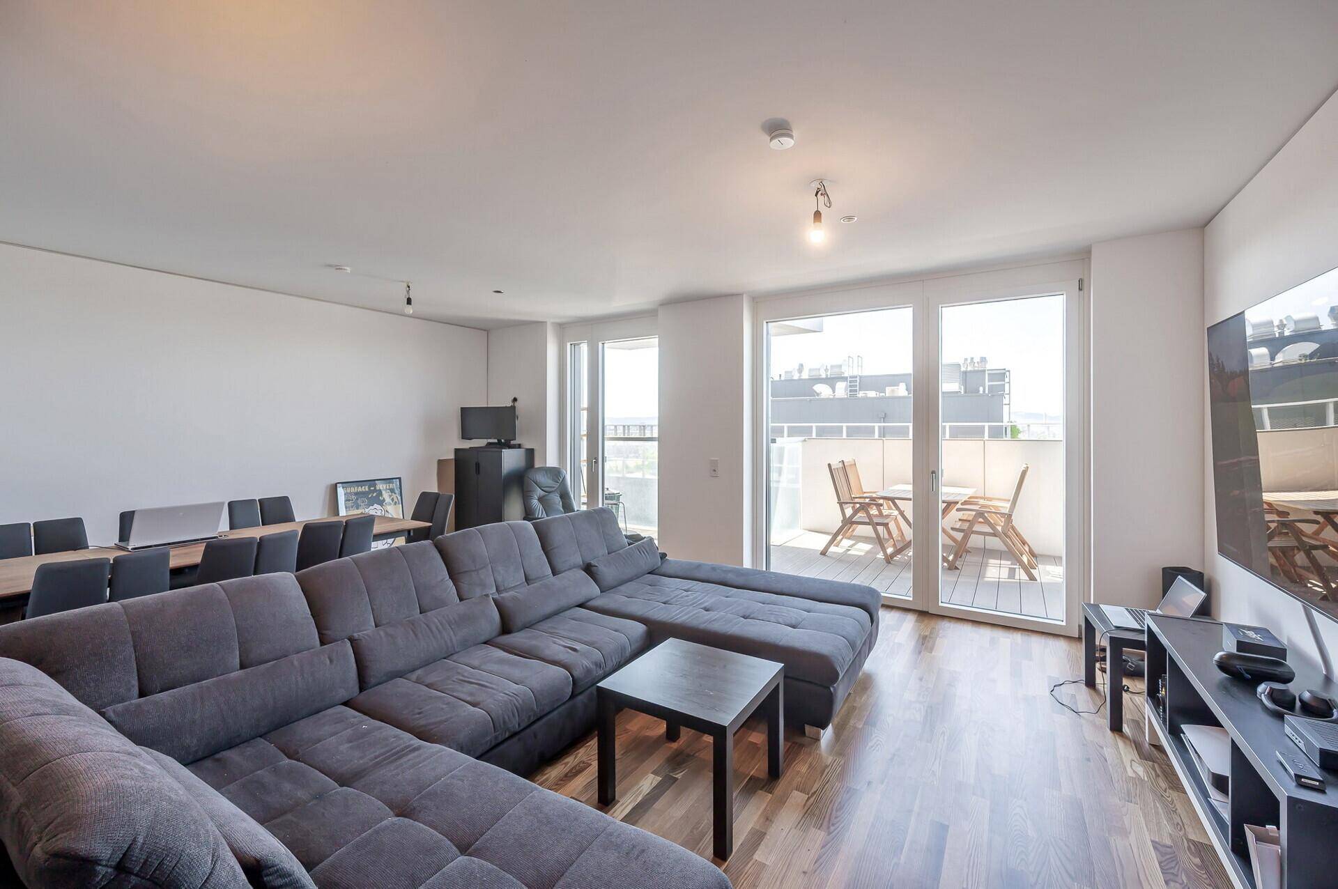 Offener Wohn- und Essbereich mit Holzboden, großem Sofa und Zugang zu einem Außenbereich.
