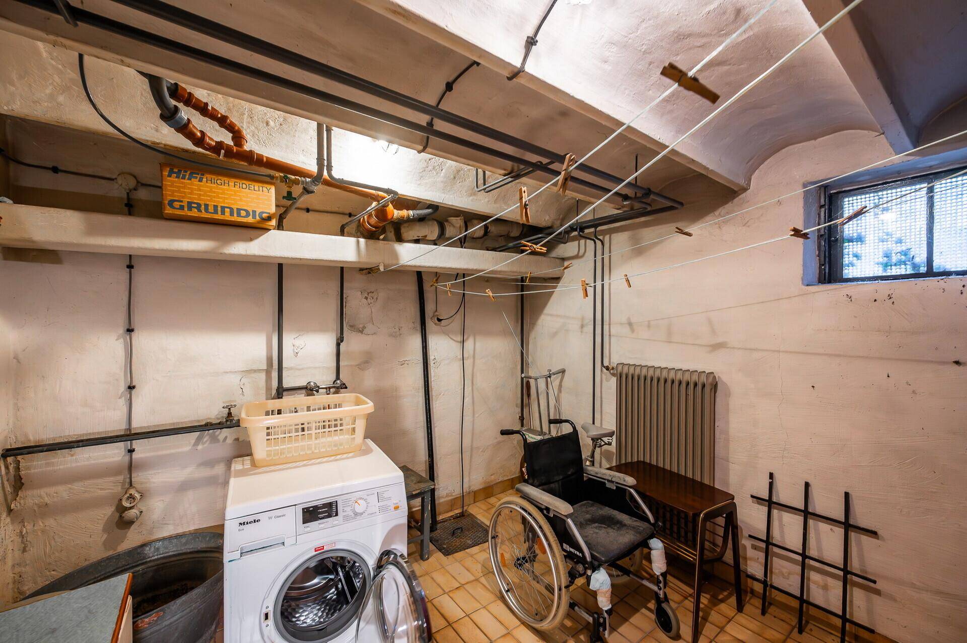 Waschküche im Keller mit Waschmaschine, Heizkörper und einem Rollstuhl, der auf Barrierefreiheit hinweist.