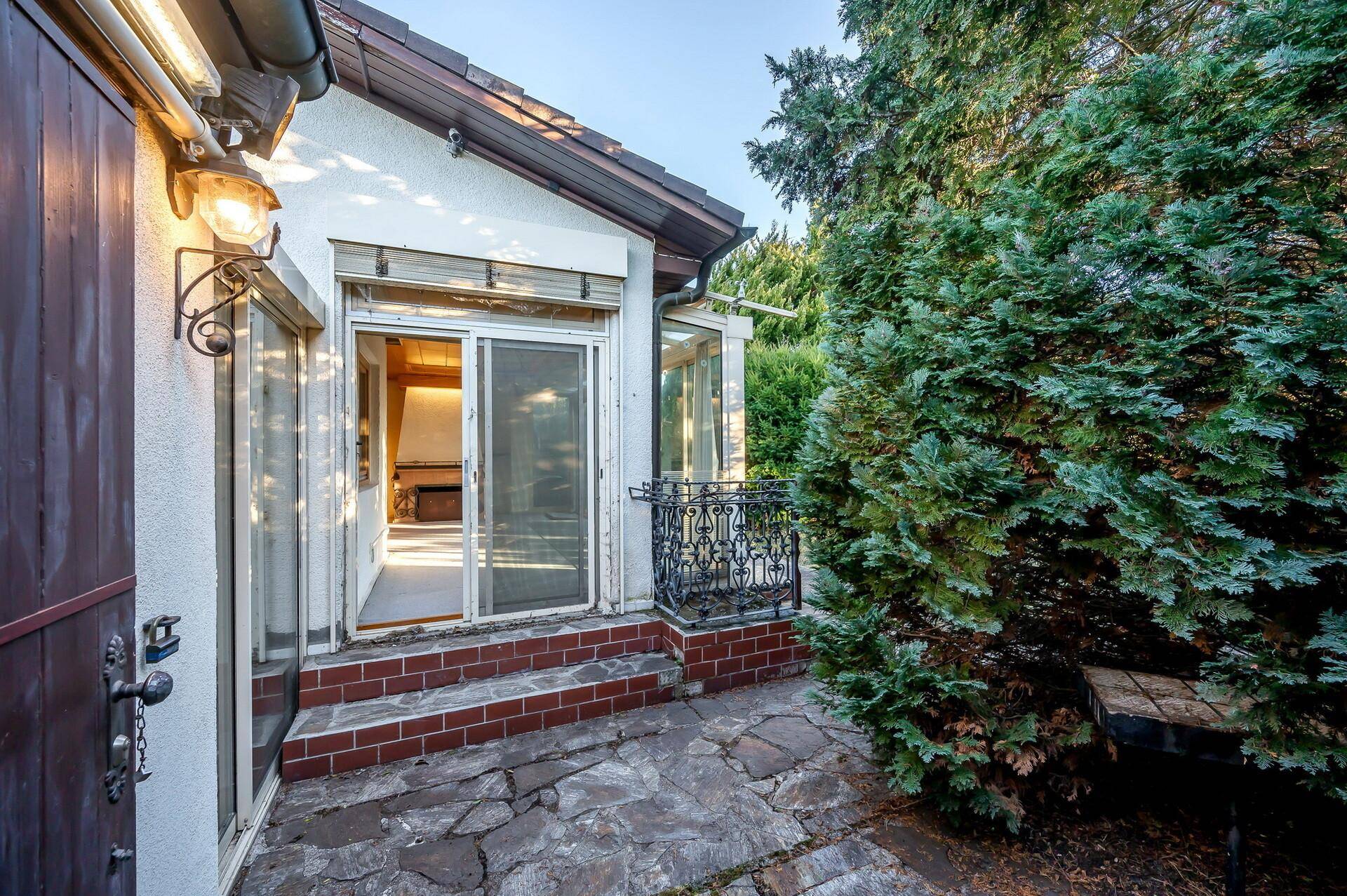 Gepflasterter Terrassenbereich mit Grillstelle und Zugang zum Wintergarten, umgeben von Grün.