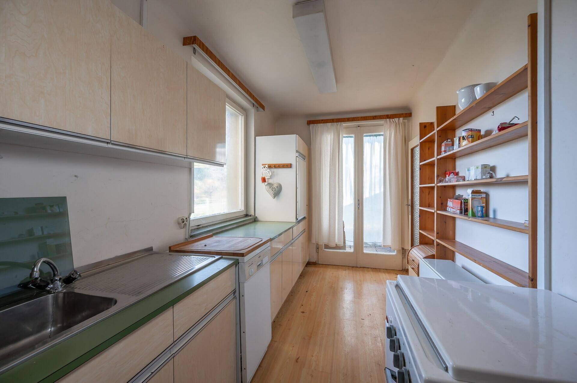 Küche mit Holzregalen, Spüle und Zugang zum Balkon oder Garten, renovierungsbedürftig.