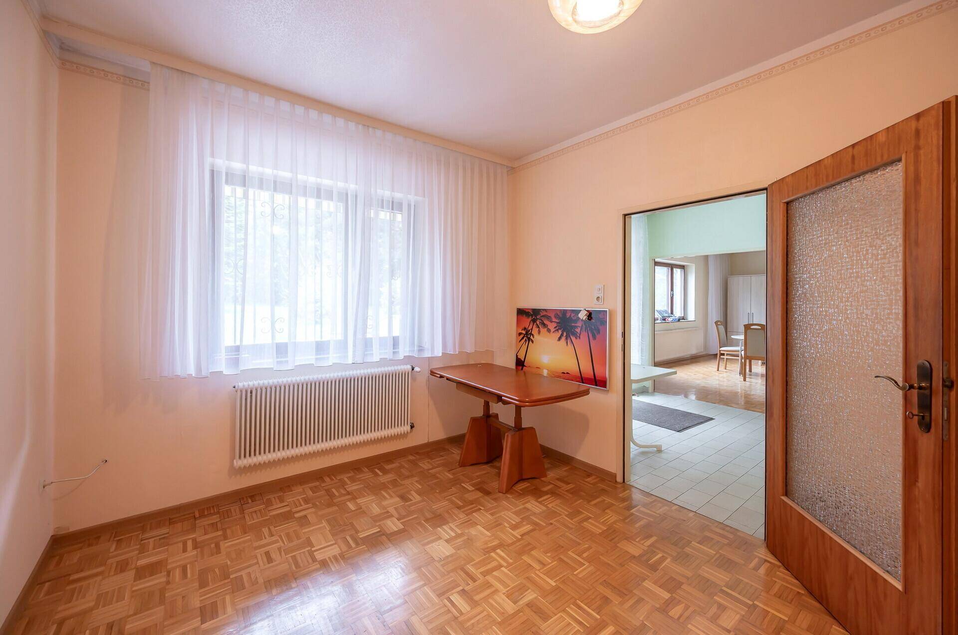 Helles Zimmer mit Parkettboden, großem Fenster und Blick in den angrenzenden Wohnbereich.