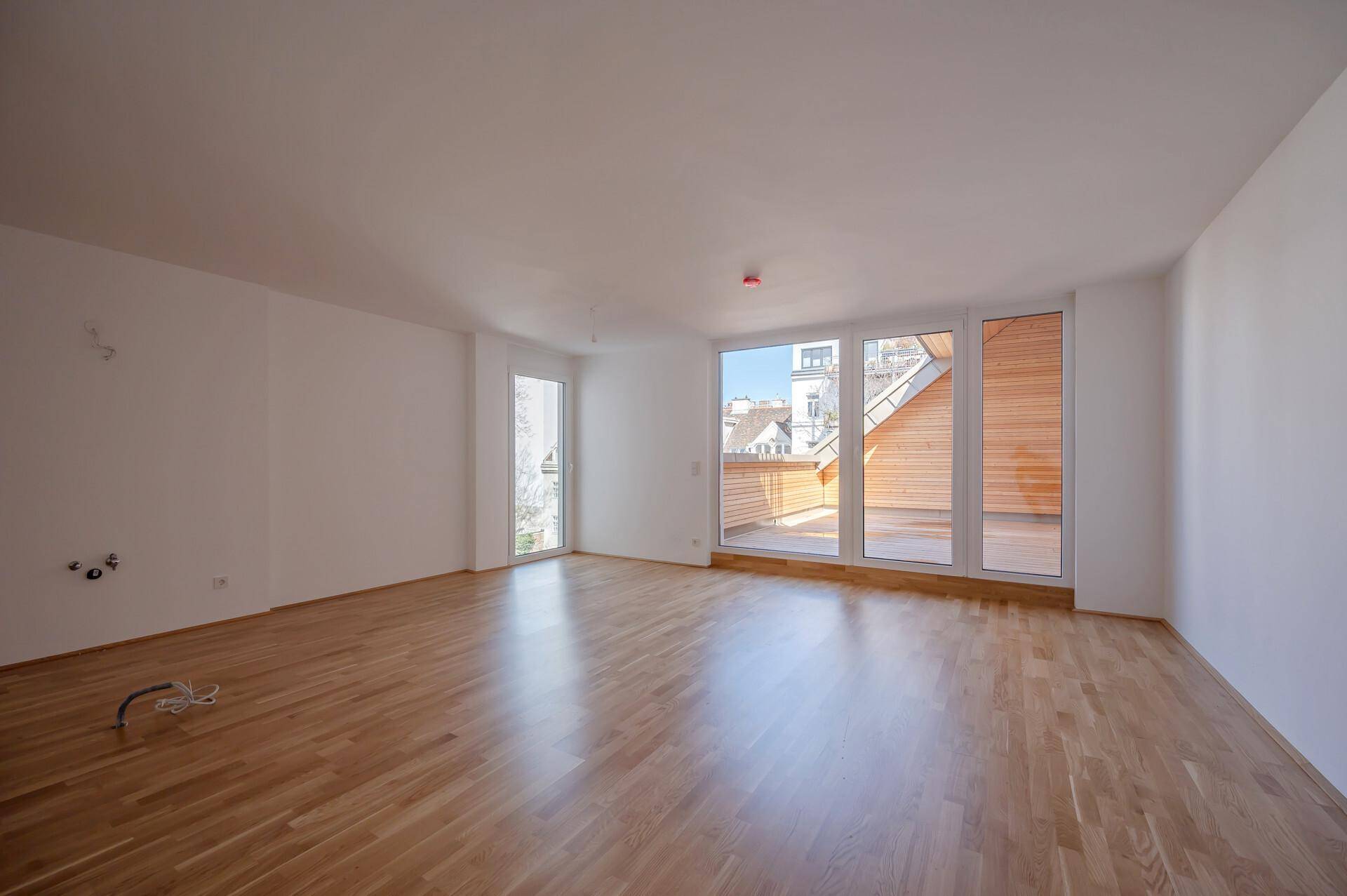 Heller, leerer Wohnraum mit Holzboden und großen Fenstern, die viel Tageslicht hereinlassen.