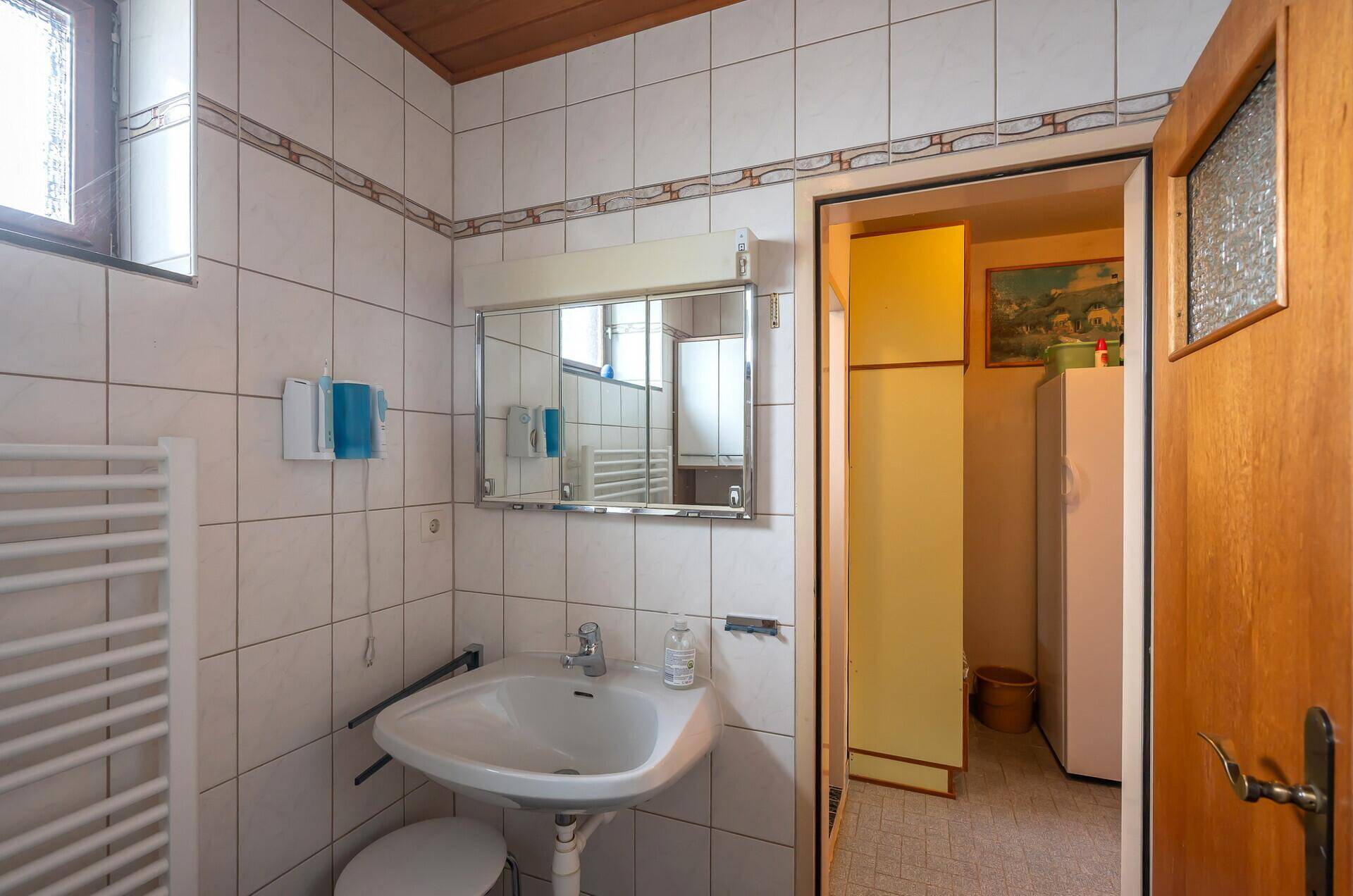 Badezimmer mit Waschbecken, Spiegelschrank und weißen Fliesen, angrenzend an einen weiteren Raum.